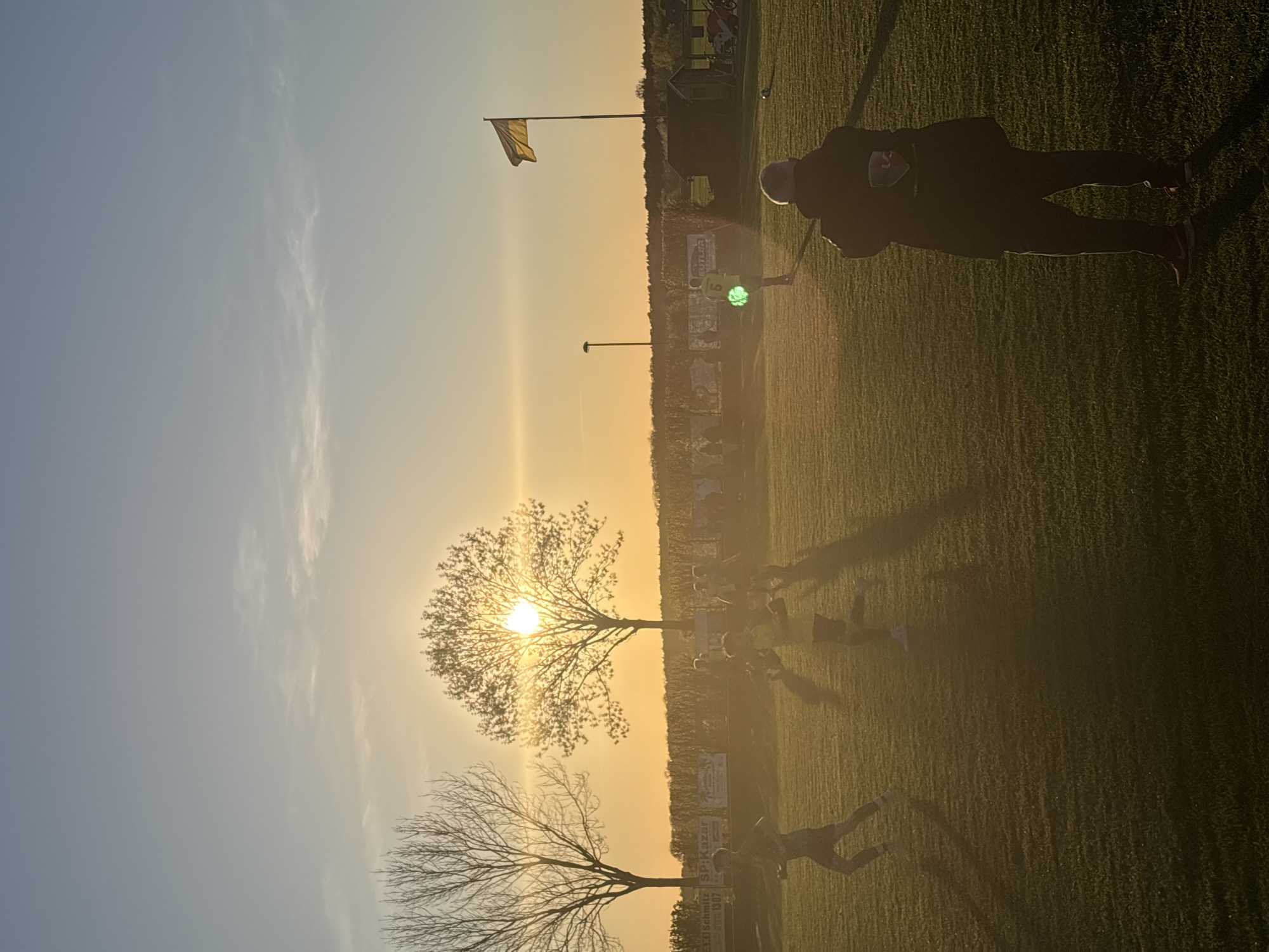 The image shows a sports field in Wegberg, Germany, at sunset. Two bare trees and one with leaves are silhouetted against the sun. A flagpole with a yellow flag is visible. Several people are on the field, including one person holding a green object in their hand. A person in dark clothing stands to the right side of the image. Advertisements are visible in the background.