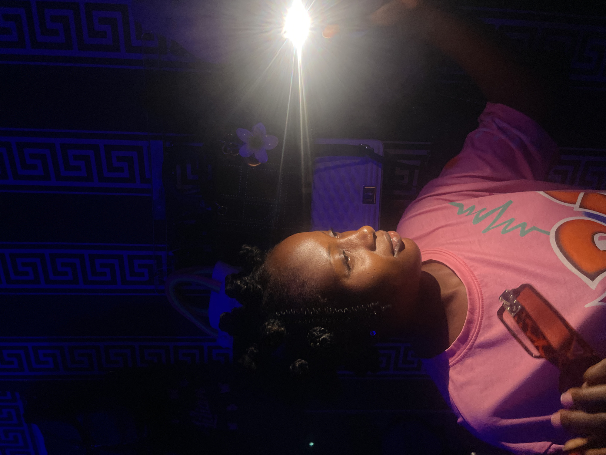 A woman is visible, holding a rectangular object near her chest. She is wearing a pink shirt with a heart design. Her hair is styled in small knots. The background consists of a dark surface with a repeating Greek key pattern. A bright light source is present above her head. The location appears to be indoors. The location in the city of Lokoja, Nigeria cannot be determined from the image.