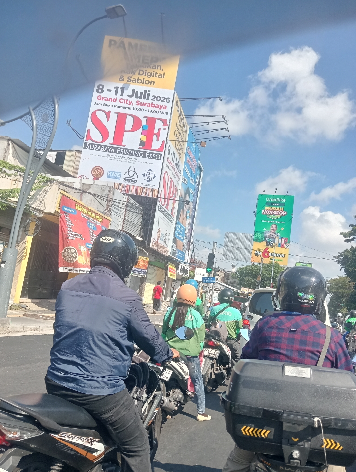 The image shows a street scene in Surabaya, Indonesia, with several motorcycles and riders in the foreground. A large billboard advertising the "Surabaya Printing Expo 2026" is visible in the background, along with another billboard for GrabBike. The sky is blue with some clouds. Traffic appears to be moderate, with several vehicles visible on the road. The overall atmosphere is that of a typical urban environment.