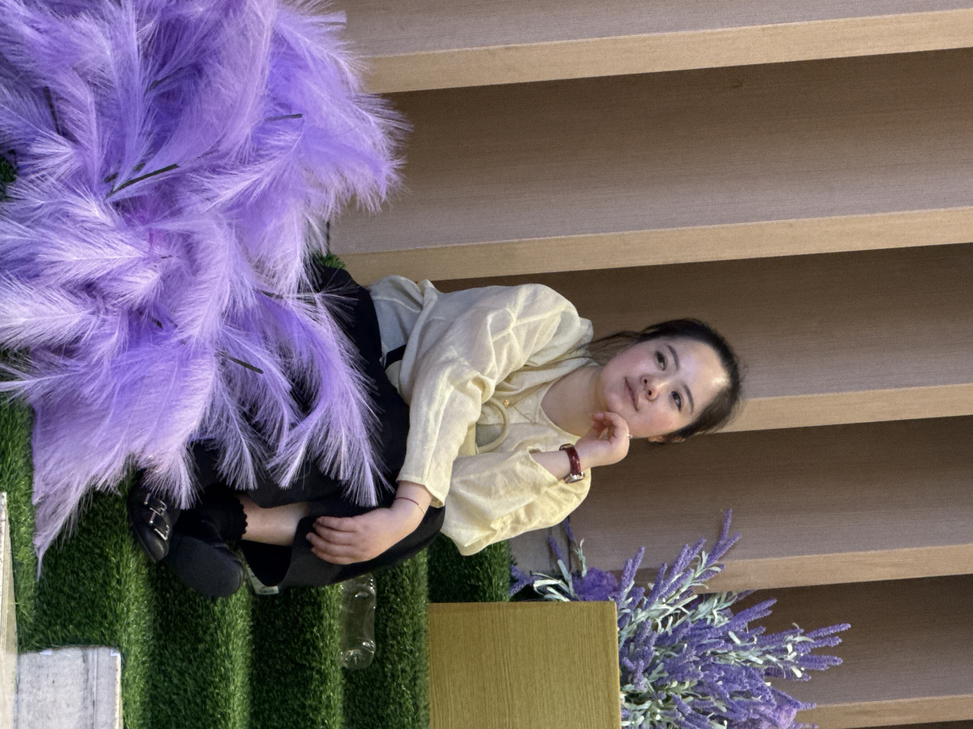 A woman sits among artificial lavender, wearing a beige top, black pants, and black shoes. She rests her chin on her hand and looks at the camera. The artificial lavender is positioned near a faux grass surface and a gold cube structure. In the background, there is a wall of light brown planks. The image appears to be taken at a location in Yuzhong District, China, based on the background.