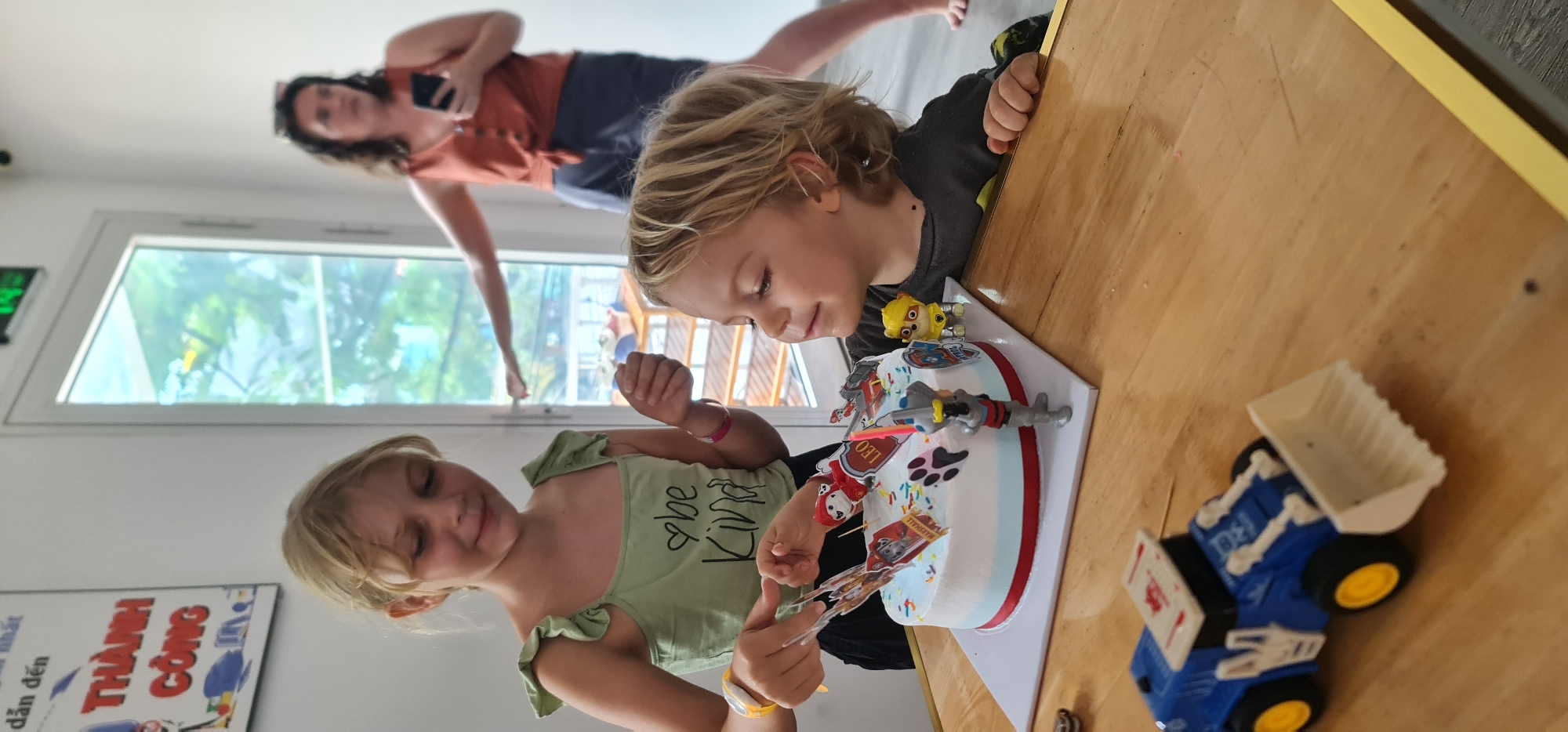 A medium, high-angle indoor shot shows two children at a table with a Paw Patrol-themed cake, while a woman is visible in the background near a window. The location is identified by a sign as being in Đà Nẵng, Vietnam.
A blond girl in a green “Be Kind” shirt sits on the left side of the frame, holding Paw Patrol-themed cake toppers. Next to her on the right is a blond boy in a dark-colored shirt, looking at the cake. The cake is decorated with Paw Patrol characters and has a red and white border. A blue and yellow toy truck with a white bed sits on the wooden table next to the cake.
In the background, a woman in an orange shirt and dark shorts stands near a window. The window offers a glimpse of foliage and an exterior structure. A sign is partially visible on the wall behind the girl in the green shirt. The setting is brightly lit, suggesting a casual, informal environment.