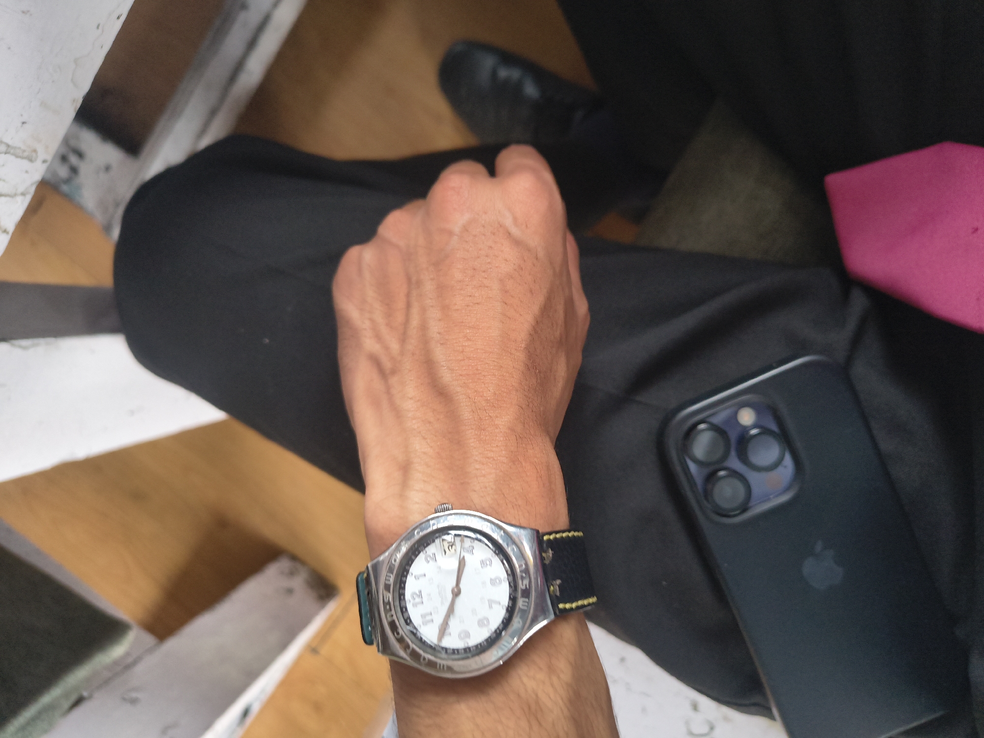 A close-up, high-angle shot shows a person's right wrist wearing a silver Swatch watch with a white face and black strap. The watch has Arabic numerals and a date window. The person's hand is resting on their lap, wearing black pants. To the left of the hand, a black iPhone with a prominent camera bump and an Apple logo is visible. In the background, parts of what appear to be wooden seating and a blurred shoe suggest an indoor setting. There is no identifiable information to place this image in Dharamshala, India.