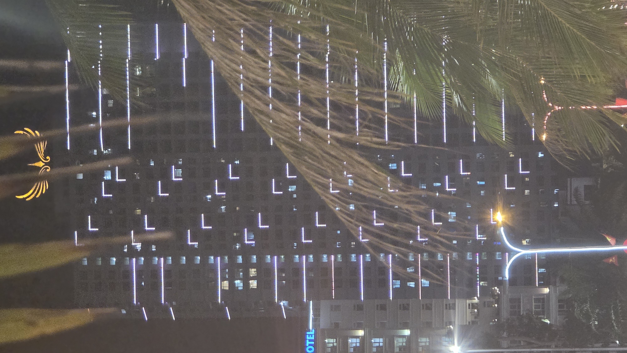 Captured at night, the photograph presents a cityscape in Nha Trang Ward, Vietnam. The main subject is a multi-story building decorated with illuminated lights. Some of the lights are vertical, while others are in the shape of the letter "L", outlining the building's structure. A palm tree partially obscures the view of the building, its fronds visible in the upper right portion of the image. The lower portion of the image shows other buildings and light sources, suggesting an urban environment. The sky is dark, and the overall light level is low.