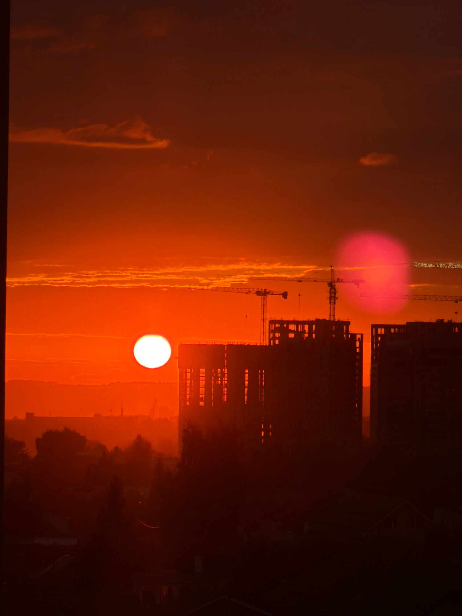 An extreme wide shot captures a sunset in Kazan, Russia, painting the sky in vivid shades of orange and red. The sun, a bright white disk, hangs low on the horizon, casting long shadows and silhouetting the cityscape. In the foreground, dark outlines of buildings and trees create a stark contrast against the fiery sky. Construction cranes are visible, suggesting ongoing development. A pinkish-red lens flare, likely from the camera, adds an unusual element to the scene, located to the right of the sun. Text, possibly a watermark or inscription, reads "Казань. Ты. Люблю." which translates to "Kazan. You. Love."