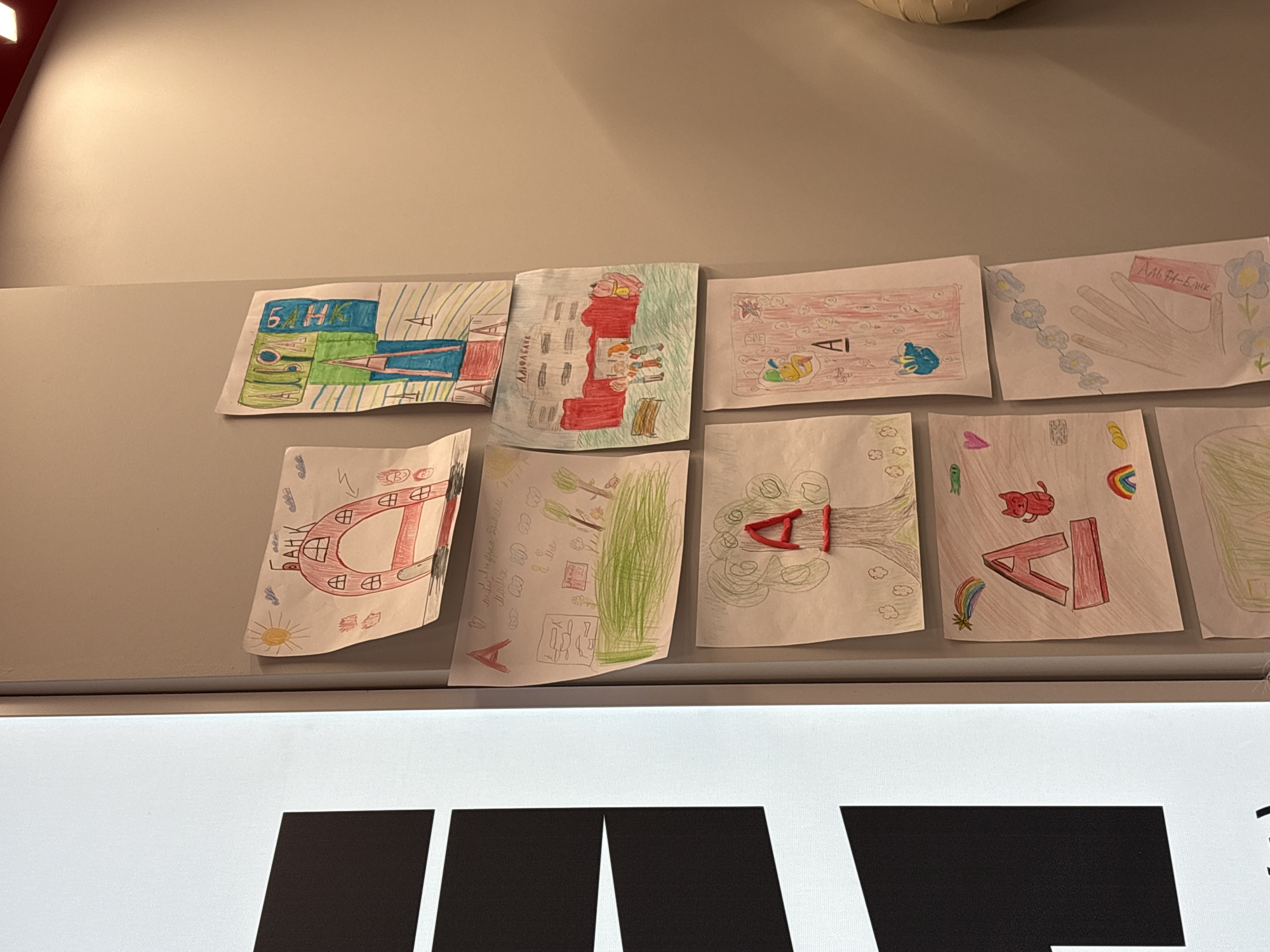 The image displays a series of children's drawings attached to a wall. The drawings appear to be themed around the Russian banking institution "Alfa-Bank" (АЛЬФА-БАНК). The drawings feature images related to the bank, such as buildings with the bank's name, and letters with the bank's name.
While the image features content related to "Alfa-Bank" which has locations in Russia, including Birobidzhan, the image alone does not explicitly confirm that the photo was taken in Birobidzhan, Russia.