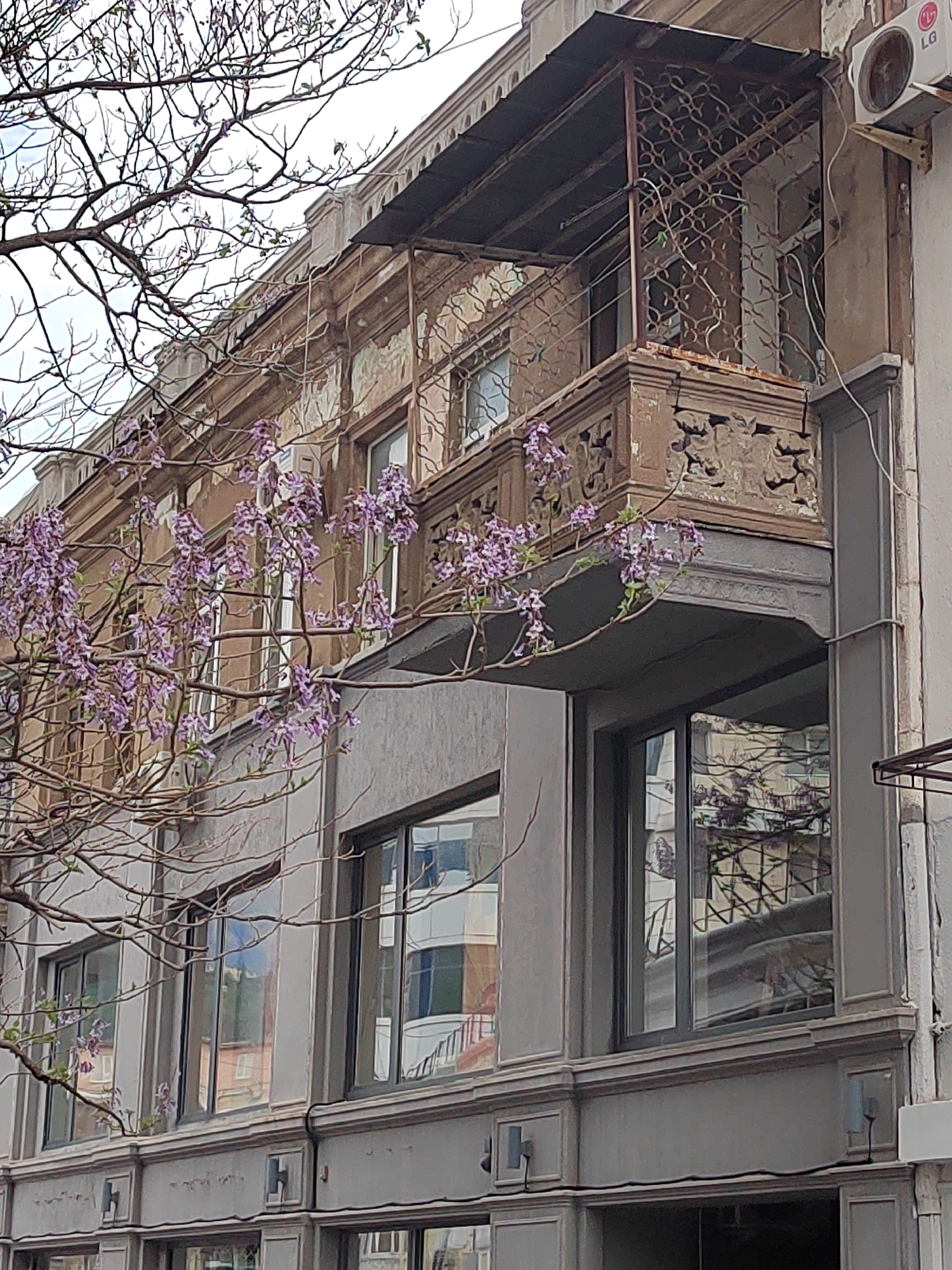 A view of a building in Unknown, Unknown. The building has a balcony with decorative metalwork. A tree with purple flowers is visible in the foreground, partially obscuring the view of the building. The building has a mix of architectural styles, with elements of classical and modern design. An air conditioning unit is visible on the side of the building. The sky is overcast.