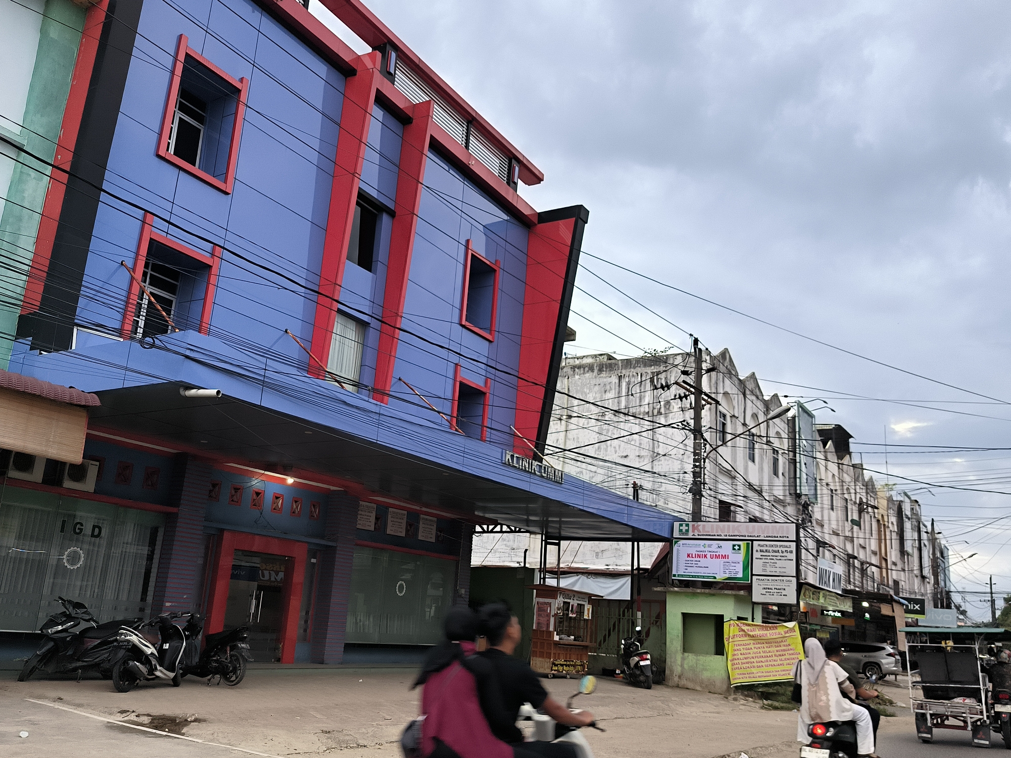 The image depicts a street scene in Langsa, Indonesia. A brightly colored, blue and red, two-story building dominates the left side of the frame. Signage on the building reads "KLINIK UMMI" and "IGD". Several motorcycles are parked in front of the building. In the foreground, two motorcycles are in motion, one with two occupants, the other with one. A white-clad woman is riding on the back of one motorcycle, while another motorcycle-drawn cart carrying a passenger is on the far right. Behind the blue and red building is a row of older, multi-story buildings, uniformly white with sawtooth roofs. Above these buildings is an overcast sky. Various signs and banners are visible along the street, displaying different businesses and information, including medical practices and local advertisements. Electrical wires crisscross the sky.
