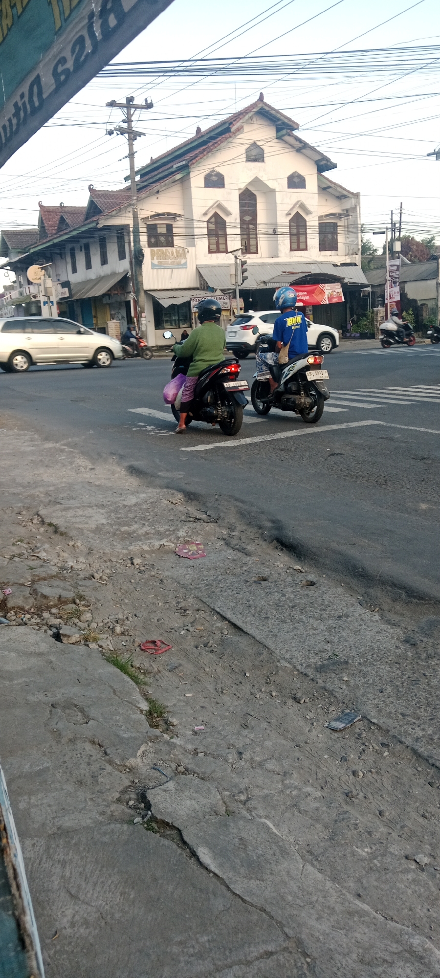 The image depicts a street scene in Maguwoharjo, Indonesia. In the foreground, the cracked and weathered concrete pavement lines the side of the road. Beyond the pavement, a paved road surface with visible crosswalk markings is visible. Two individuals are present, each riding a motorcycle. The first individual, on the left, is an elderly woman wearing a green long-sleeved shirt and patterned pants. She is on a dark-colored motorcycle with a purple bag slung over the back. The second individual, on the right, is a younger person wearing a blue shirt and a helmet, riding a lighter-colored motorcycle. In the background, several buildings are visible, including a white building with a distinct roofline and arched windows. Various utility poles and wires crisscross the sky above the street. The scene suggests an urban or semi-urban environment, likely during daylight hours, given the lighting conditions.