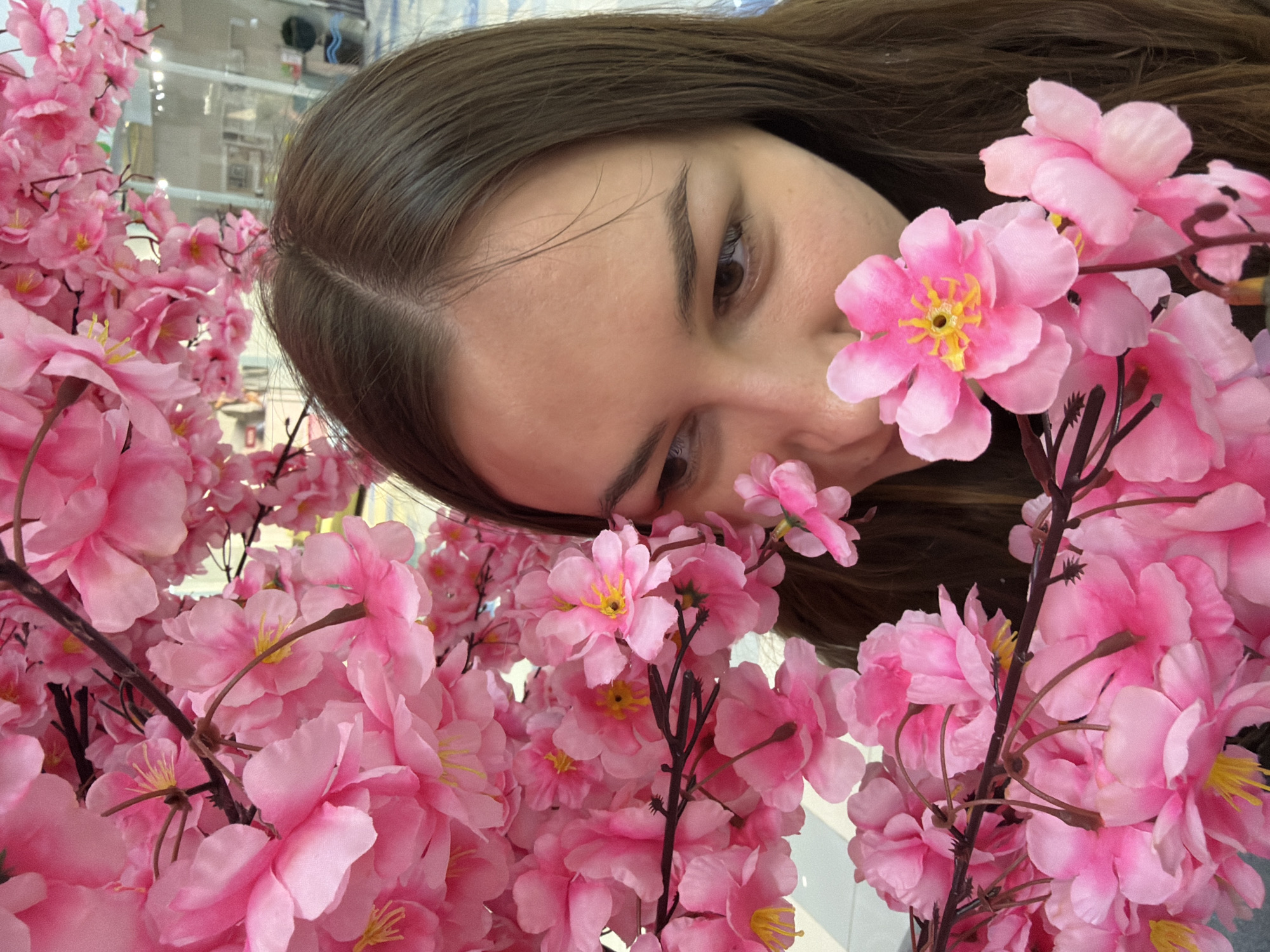 The image captures a woman with brown hair, gazing down with a soft focus. Her face is framed by an abundance of pink cherry blossom flowers, both real and artificial. The flowers are densely packed around her, with the stems and branches visible in the mix. The location is likely a commercial setting, judging by the shelving and interior details visible in the background, and it's in the city of Izhevsk, Russia.