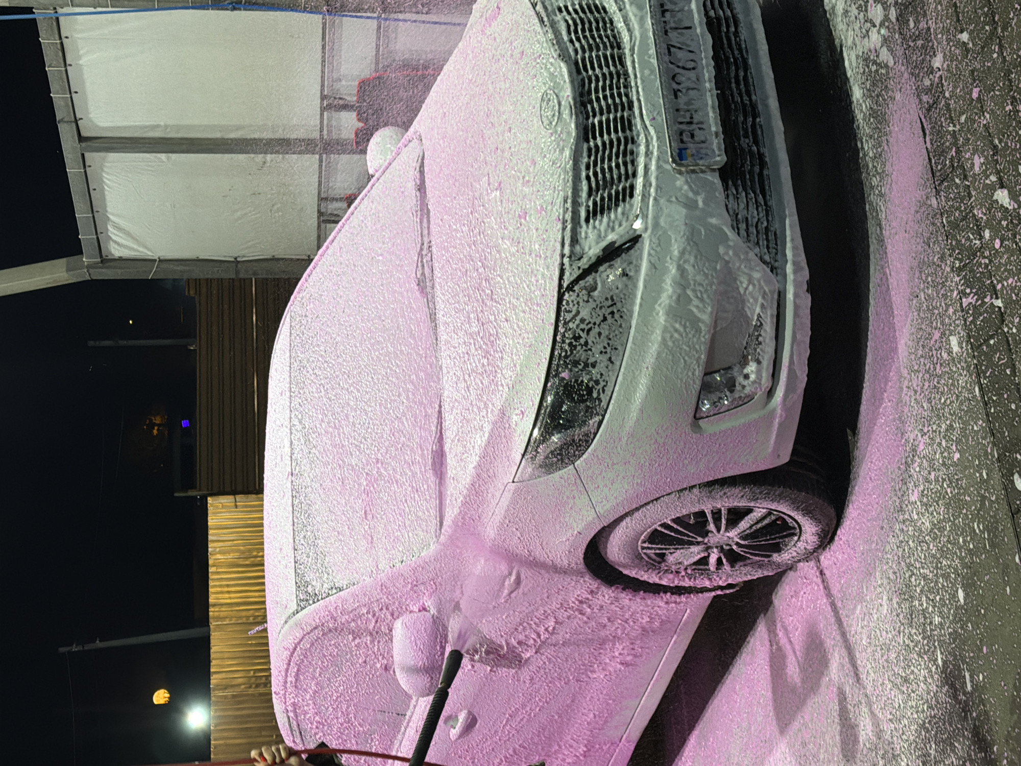 A white car is being washed with pink foam at a car wash in Korsuntsi, Ukraine. The car is covered in a thick layer of foam, and the ground around it is also covered in foam. A person's hand is visible on the left side of the image, holding a spray wand that is spraying the foam onto the car. The car wash appears to be outdoors, with a fence and trees visible in the background. The lighting suggests it is nighttime.