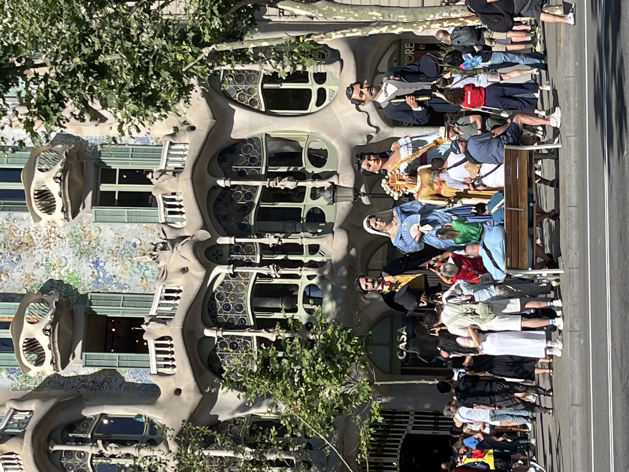 This is a full, eye-level, outdoor, daylight, medium-shot photograph of Casa Batlló, a famous building located at Passeig de Gràcia in Barcelona, Spain. The building features a distinctive, organic facade with undulating shapes and vibrant mosaic tiles. In front of the building, there are three oversized, traditional Catalan figures, known as Gigants, which are commonly seen in festivals and celebrations. The figures are dressed in elaborate costumes: one in a blue dress and headdress holding a fan, another in a golden dress and ornate crown, and a third in a dark suit and top hat. Between the Gigants, a wooden bench is visible, with several people sitting and standing nearby. The foreground shows a sidewalk and a street, with a diverse group of tourists and locals gathered, many of whom are taking pictures of the building and the Gigants. Trees with green foliage are present on the right side of the frame, partially obscuring the building. The sky is clear and blue. The atmosphere appears to be lively and touristic.