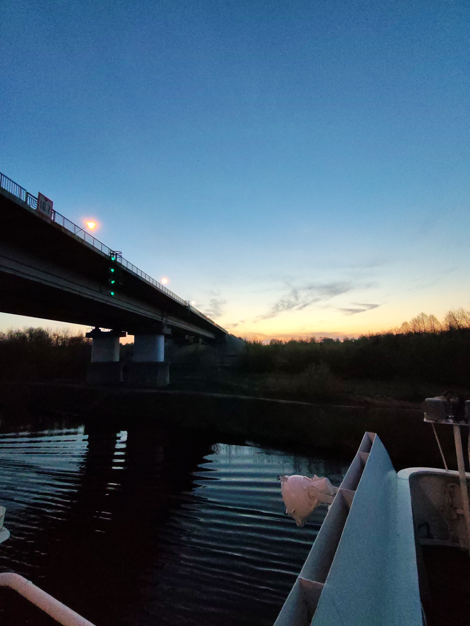 The image shows a view from a boat on a river, with a bridge in the background. The sky is a mix of blue and orange, indicating dusk or dawn. The river reflects the colors of the sky. The bridge is made of concrete and steel, with lights illuminating its structure. The foreground includes parts of the boat, such as a railing and some equipment. The shoreline is visible with trees and vegetation. The location is Unknown, Unknown.