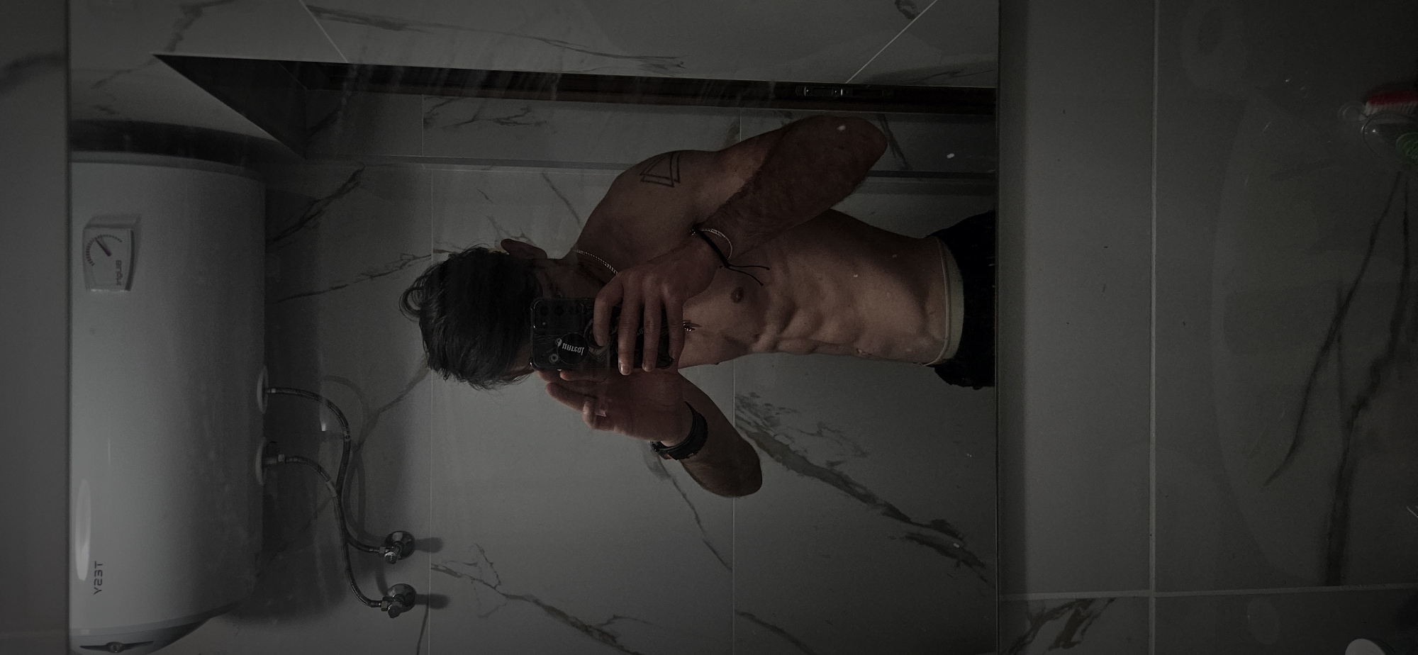The image shows a shirtless man taking a selfie in a bathroom. The man is reflected in a mirror. He is holding a black smartphone. He has dark hair and appears to be wearing dark shorts. The bathroom has marble-patterned tiles, a water heater on the left, and a shelf at the top of the mirror. The location is not identifiable as Plovdiv, Bulgaria, based solely on the image.