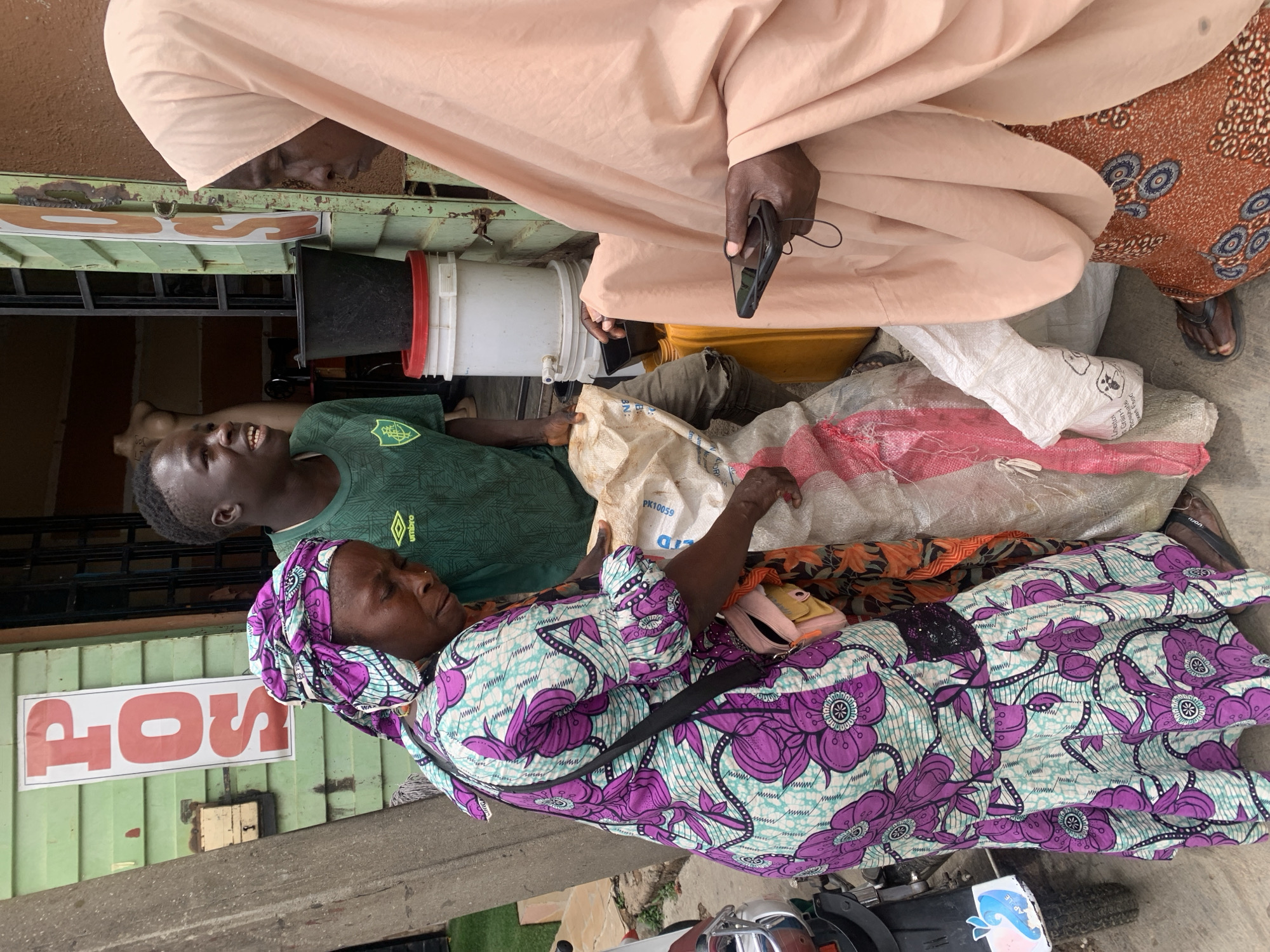 The image shows a street scene in Jos, Nigeria. A woman in a purple and white floral print dress and headwrap lies on a pile of sacks, near what appears to be a motorcycle. A young man in a green soccer jersey stands nearby, looking up and to the left. Overhead, an individual in a peach-colored robe is holding a mobile phone. Another person wearing a brown and orange patterned fabric stands near the robed person. Buckets and containers are visible behind the young man. A sign reading "POS" is affixed to a green wall in the background.