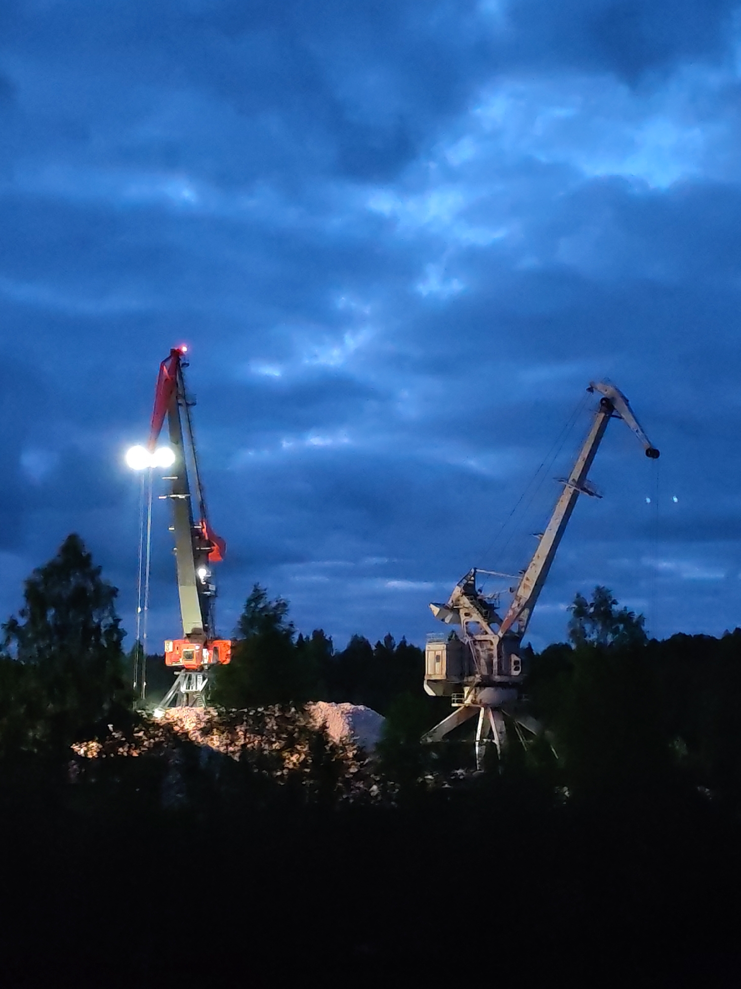 Two cranes are situated amidst trees under a dark, cloudy sky in Александровское, Russia. The crane on the left is red and grey with lit lights. The crane on the right is grey. Below the cranes is a pile of materials.