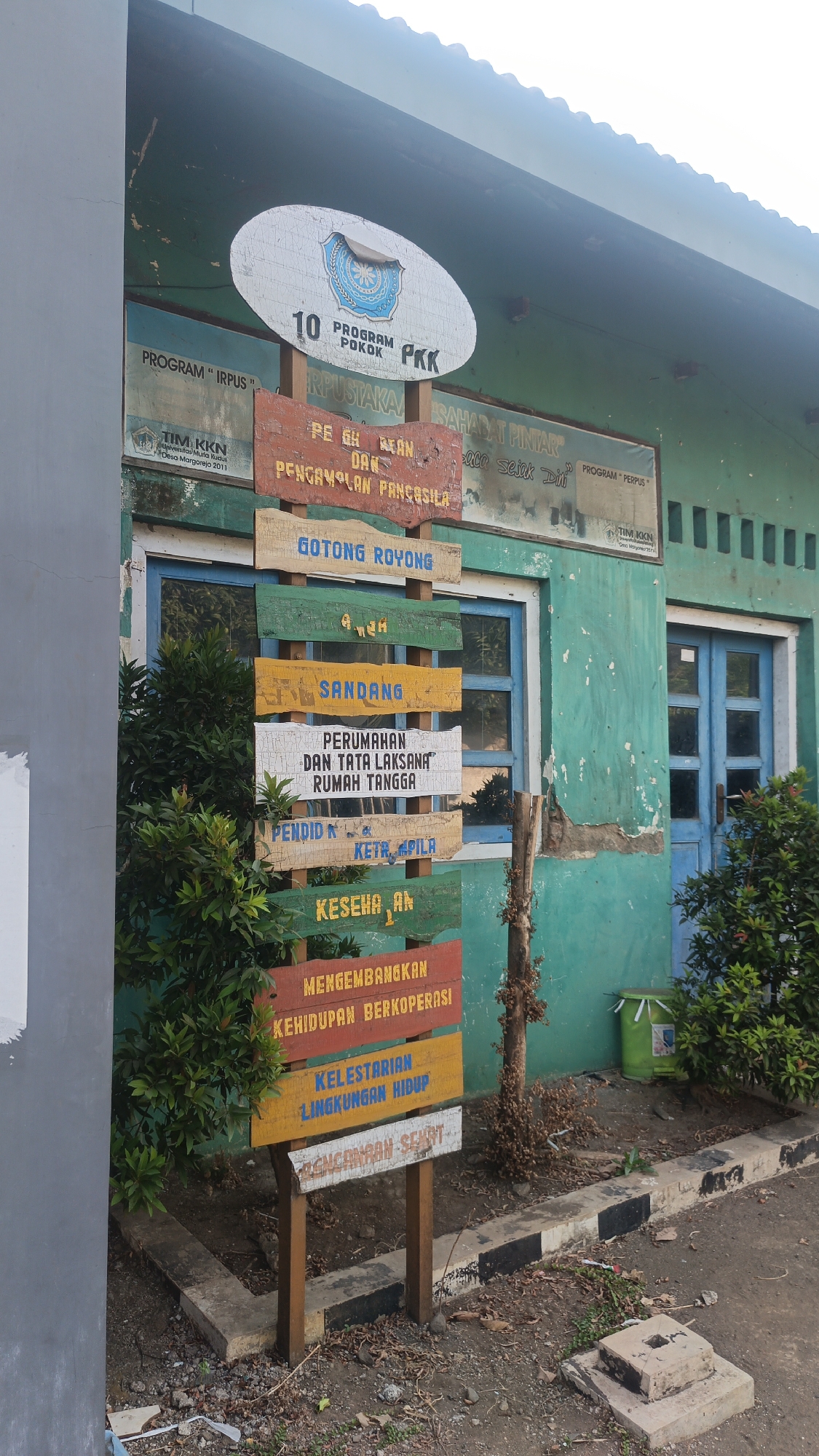 The image shows a signpost with multiple wooden planks of various colors, each displaying text in Indonesian. The signpost is located outside a teal-colored building with blue-framed windows and a blue door. Green foliage is visible on both the left and right sides of the signpost, with the building's foundation at the bottom. Debris and dry leaves are scattered on the ground around the signpost.
The text on the signpost can be partially deciphered. The topmost plank, which is oval, has the text "10 PROGRAM POKOK PKK" along with a logo. Below this, other planks read:
* "PROGRAM 'IRPUS'"
* "PE GH AT AN DAN PANCASILA" (partially obscured)
* "GATONG ROYONG"
* "SANDANG"
* "PERUMAHAN DAN TATA LAKSANA RUMAH TANGGA"
* "PENDIDIKAN KETRAMPILAN" (partially obscured)
* "KESEHATAN"
* "MENGEMBANGKAN KEHIDUPAN BERKOPERASI"
* "KELESTARIAN LINGKUNGAN HIDUP"
* "PERENCANAAN SEHAT"
The building appears to be a public facility, possibly a community center or school, given the nature of the text on the sign. The location is identified as Dawe, Indonesia.