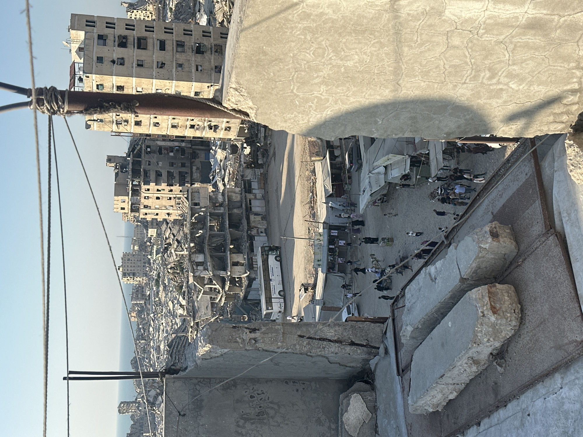 This is an aerial view of a street in the city of Gaza, Palestinian Territories, depicting extensive destruction of buildings and infrastructure. In the foreground, two large concrete blocks rest on a metal surface. The middle ground shows a street with a white bus, several cars, and numerous people walking and gathered. Some structures appear to be makeshift stalls or shops. Beyond the street, a vast expanse of destroyed buildings and rubble stretches towards the horizon, with the Mediterranean Sea visible in the distance. The overall scene conveys a sense of widespread devastation and the difficult living conditions in the area.