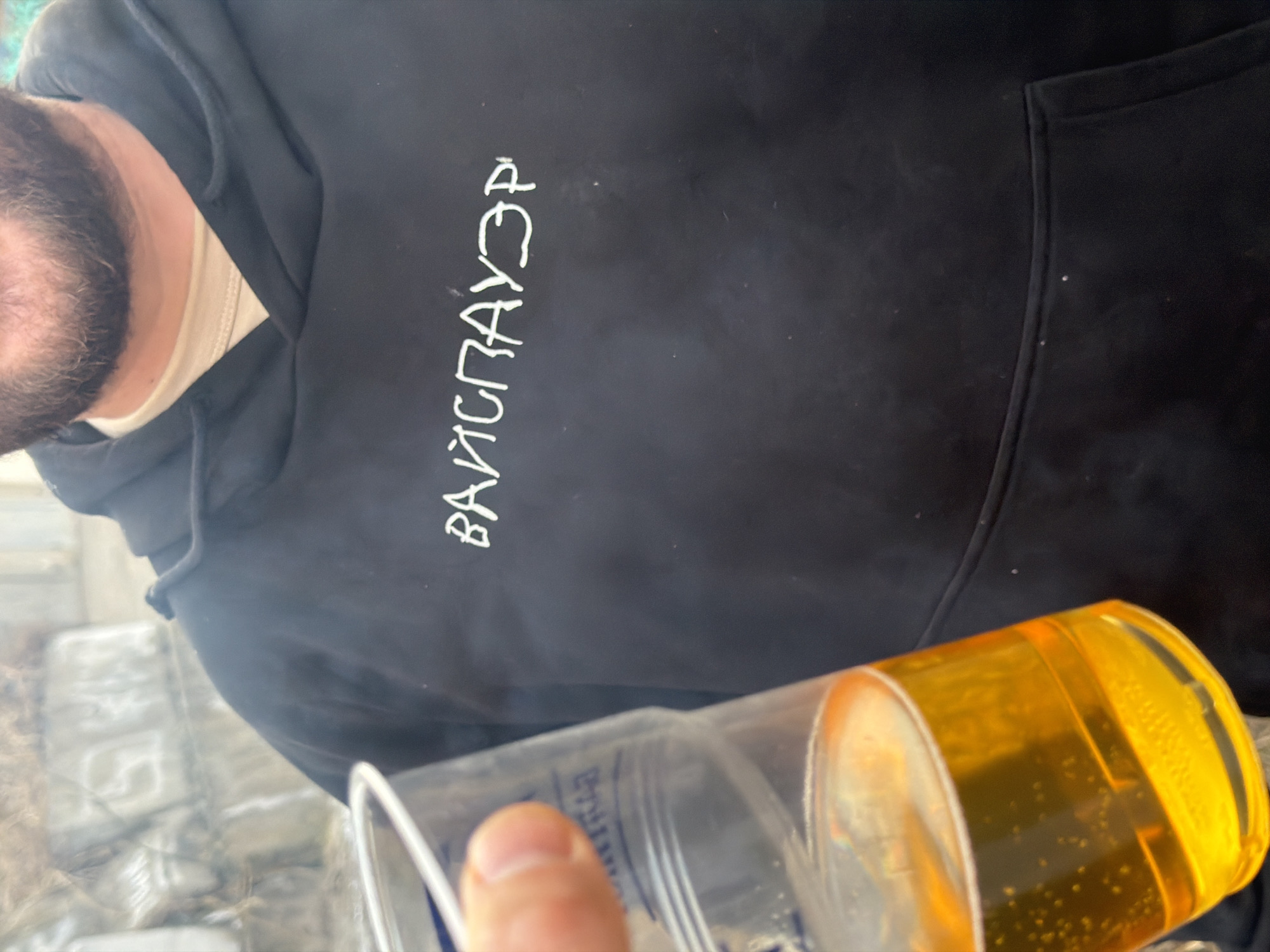A close-up shot features a person wearing a black hoodie with text written in Cyrillic ("ВАЙСПАУЭР") vertically along the chest. The person has a beard. A clear plastic cup filled with a yellow liquid, possibly beer, is in the foreground, partially obscuring the lower portion of the hoodie. The location is not definitively discernible as Krasnoyarsk, Russia, based solely on the provided image. The background shows indistinct, blurred shapes suggesting an outdoor setting with possible structures.