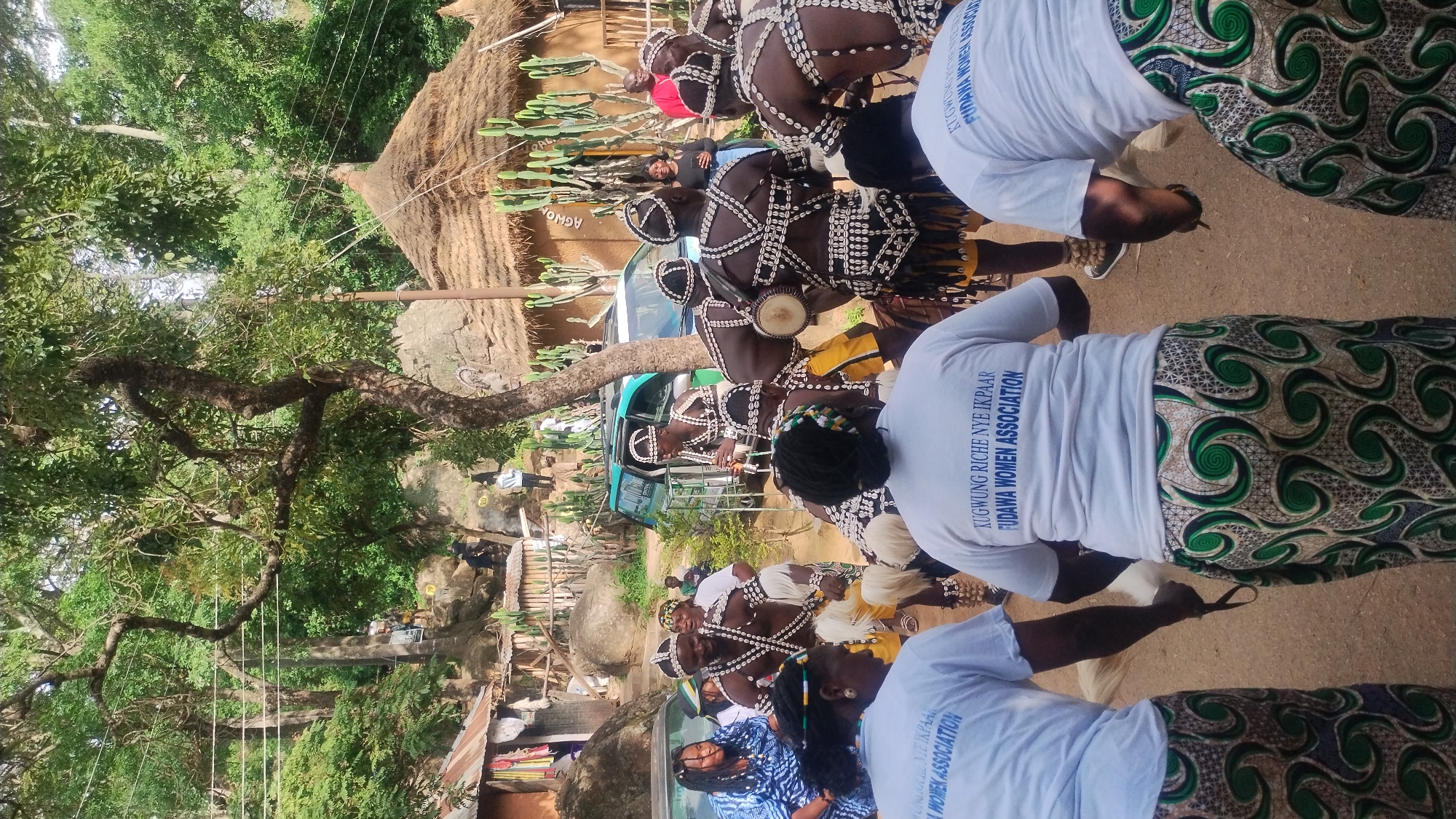 In Jos, Nigeria, a group of people are gathered in an outdoor setting that appears to be a village or cultural center. In the foreground, two women wearing white t-shirts with blue lettering that reads "KUGWUNG RICHE NYE IKPAAR FUDAWA WOMEN ASSOCIATION" and patterned skirts with green and white swirls are facing away from the camera, their backs visible. They are participating in a cultural event, as evidenced by the traditional attire worn by other individuals in the scene.
A group of men in the mid-ground are dressed in elaborate traditional costumes, featuring beaded necklaces and chest adornments made of white shells, yellow loincloths, and elaborate headdresses. Some of these men are playing instruments, including what appear to be drums. Another man in the mid-ground is playing a flute. A woman in a blue and white patterned dress is visible to the left, and other people are scattered throughout the scene, some observing the performance.
The environment includes lush green trees and vegetation, rocky outcrops, and traditional thatched-roof huts with mud walls. Two vehicles, a light blue SUV and a darker colored sedan, are parked in the background, suggesting the event may be accessible by modern transportation. The overall atmosphere is festive and celebratory, indicative of a cultural performance or gathering. The ground is covered in dirt and sand. The image is taken during the daytime, with natural light illuminating the scene.
