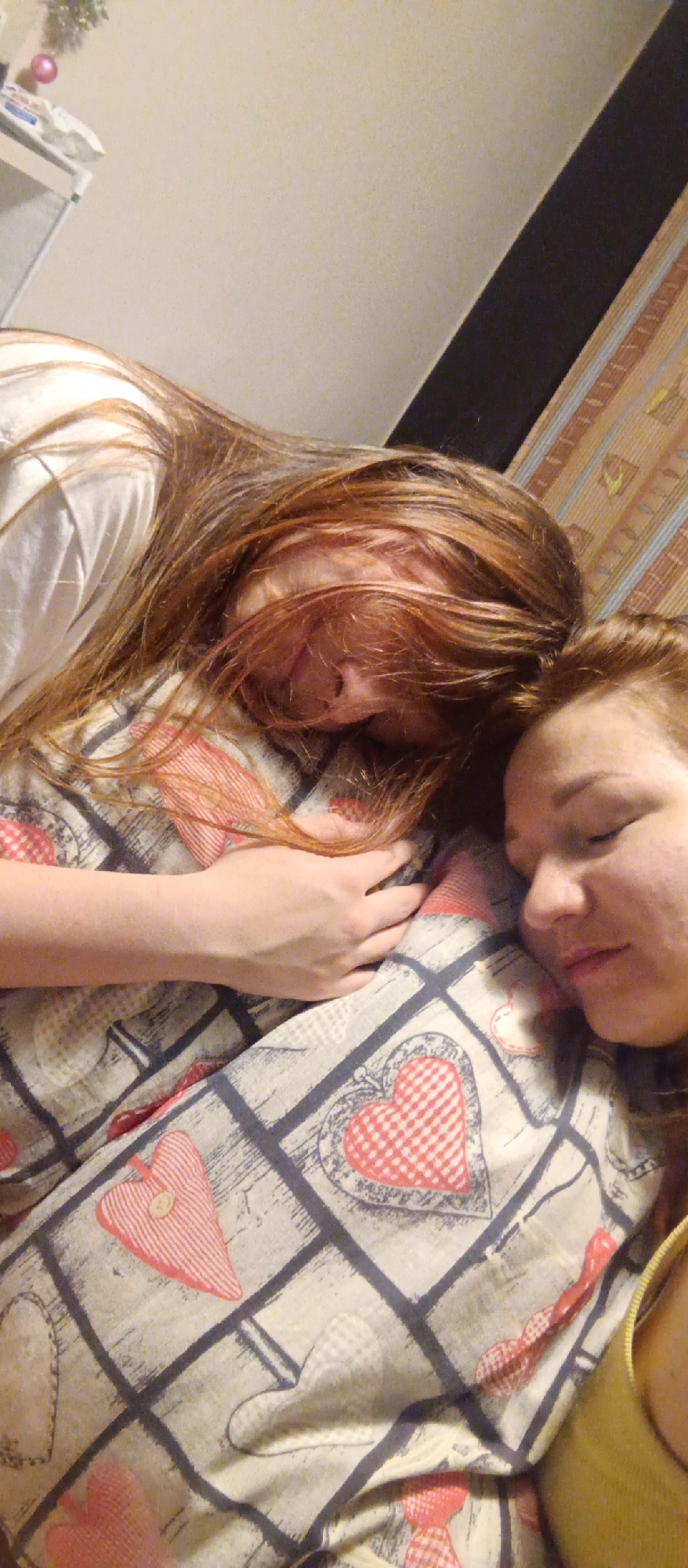 The image shows two individuals lying down, presumably sleeping. One individual has long reddish-brown hair that covers a portion of their face and hand resting on a patterned pillow. The second individual, with auburn hair, is visible from the forehead down, eyes closed. They are lying on a pillow with a grid pattern and various heart shapes. The location and city are unknown. A white shelf and a decorative element, possibly related to Christmas, are visible in the upper left corner. A dark panel and a patterned surface are visible in the upper right corner.