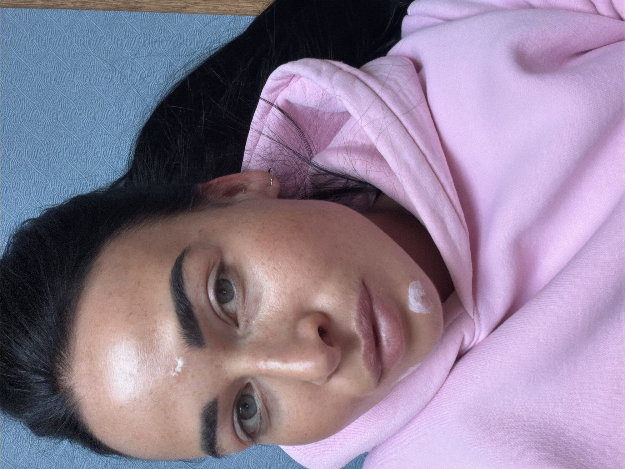 A young woman with dark hair and light eyes, wearing a pink hooded sweatshirt, is lying on a blue surface. There are spots of white cream on her face. An ear piercing is visible in her left ear. No elements in the image suggest the location is Saint Petersburg, Russia.