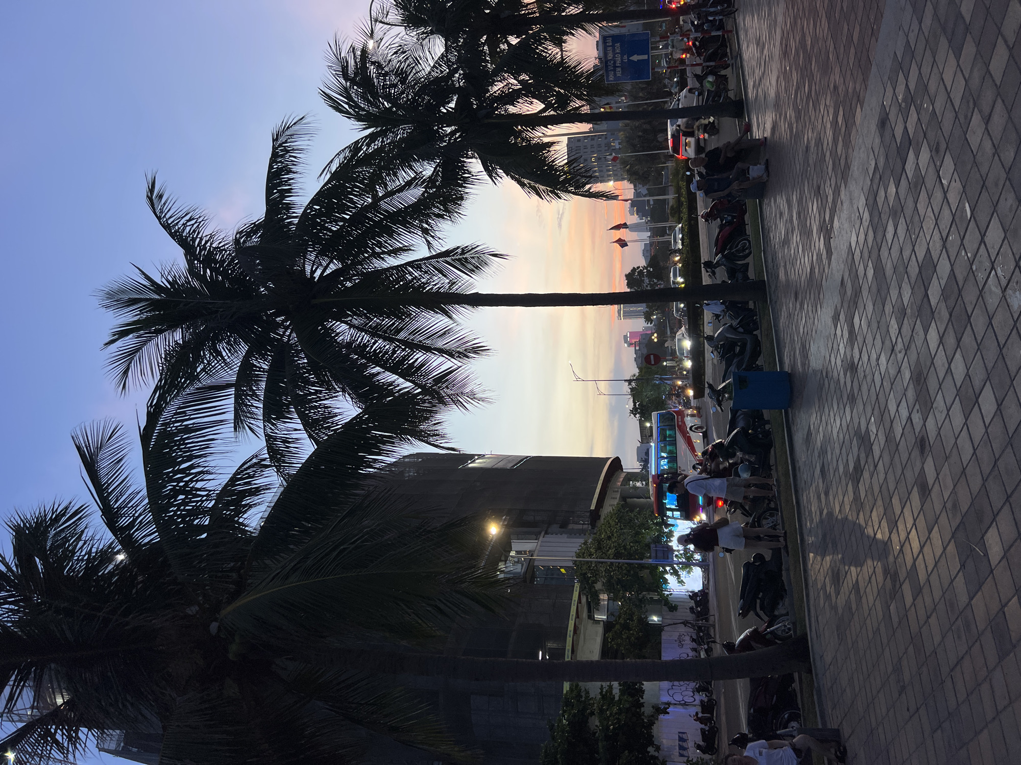 The image depicts a city street scene at dusk. In the foreground, a paved sidewalk with a tiled pattern is visible, with shadows cast by palm trees. Several parked motorcycles line the edge of the sidewalk. Further back, a road is lined with more palm trees and a building under construction, covered in black netting. A red and white bus and other vehicles are moving on the road. People are seen standing and sitting on the sidewalk. The sky displays a gradient of colors, transitioning from a pale blue near the top to hues of pink, orange, and yellow near the horizon, indicating sunset or twilight. The overall impression is a vibrant urban environment at the end of the day.