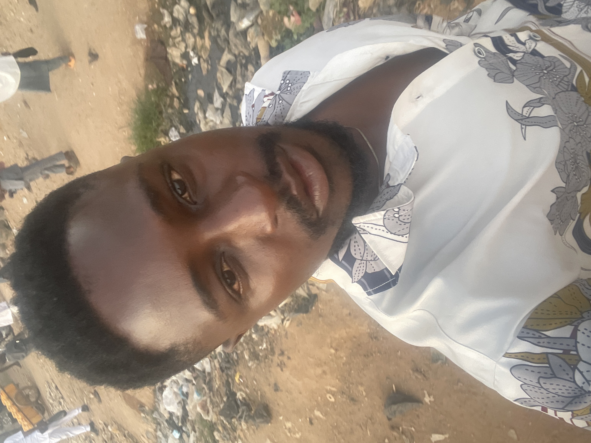 A close-up, slightly rotated image shows a man with dark hair and a trimmed beard. He is wearing a white shirt with a floral print. The background appears to be an outdoor area with gravel, dirt, and scattered debris, possibly indicating a less-developed or neglected area. In the background, blurred figures of people can be discerned. The location is in the city of Abuja, Nigeria.