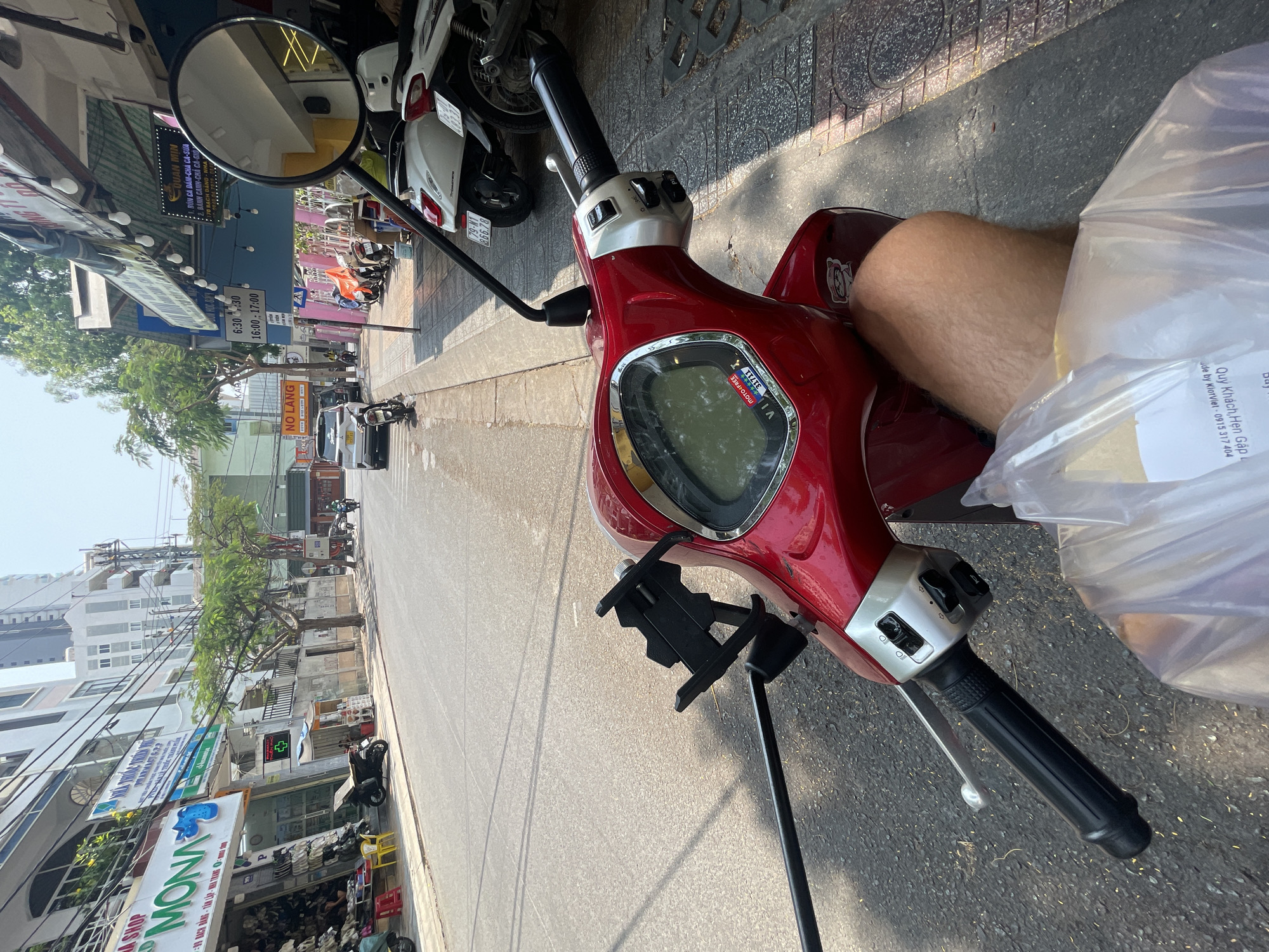 The image is a perspective shot from the seat of a red scooter, showing the view down a city street in Nha Trang, Vietnam. In the foreground, the scooter's handlebars, instrument panel, and a mobile phone holder are visible. A person's leg and a plastic bag are positioned on the right side of the scooter.
The street scene includes various shops and businesses, such as a shoe store named "MONA", a pharmacy (PHARMACYG.23), and a restaurant "QUAN MIN". Street signs and advertisements in Vietnamese are present. Other motorbikes and cars are parked or traveling along the street. The time shown on a sign is 6:30-11:30 and 16:00-17:00. Trees line the street, and buildings are visible in the background under a hazy sky.