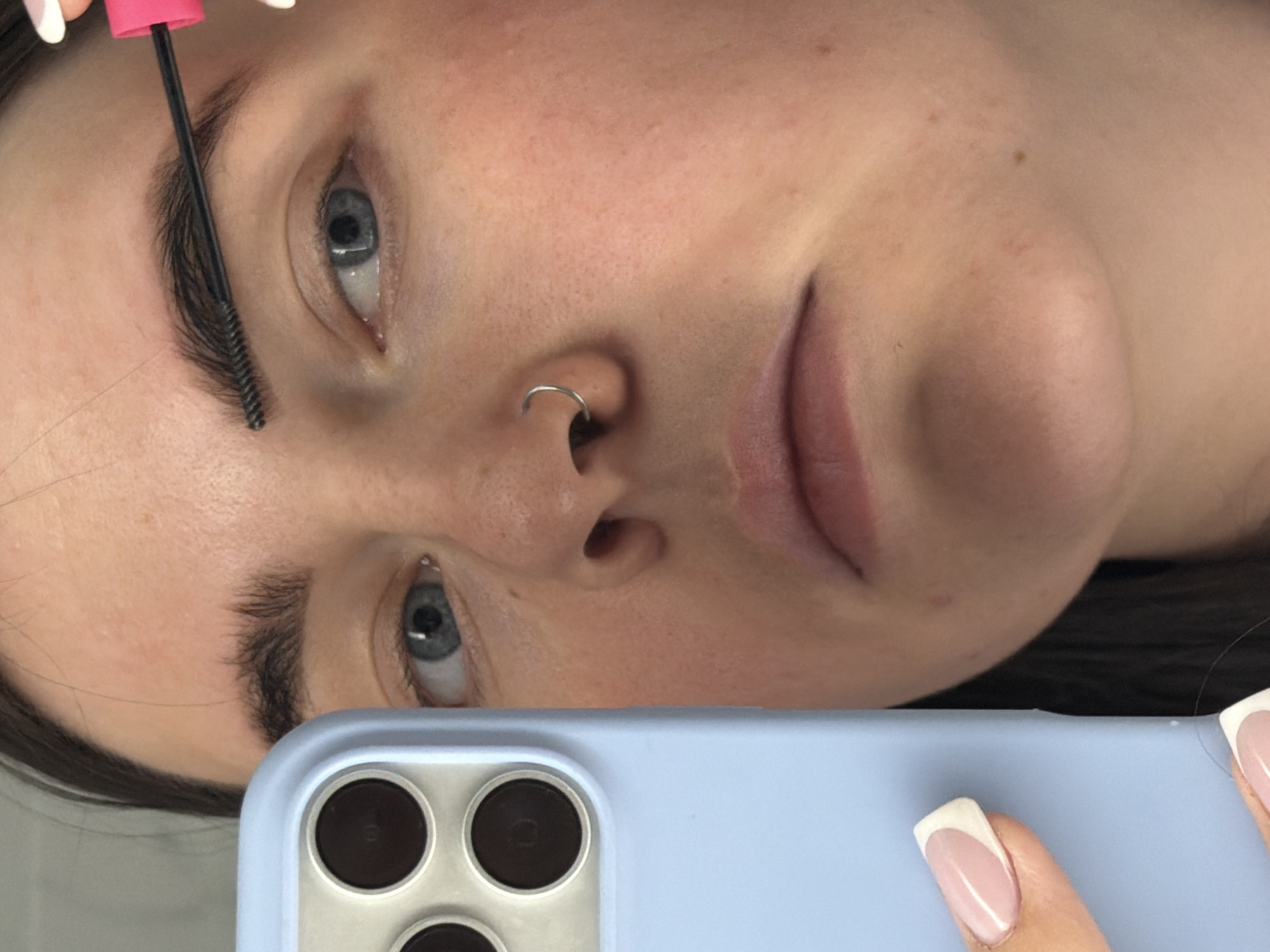 A close-up, angled shot shows a person's face, positioned at a diagonal to the viewer. The individual is holding a blue smartphone, obscuring the lower portion of their face. The person has fair skin, light blue eyes, and a nose ring. A pink-handled cosmetic brush is near their eyebrow. The person's fingernails are manicured with a French tip design. The context of the location, Chaikovsky, Russia, is not explicitly visible in the image.