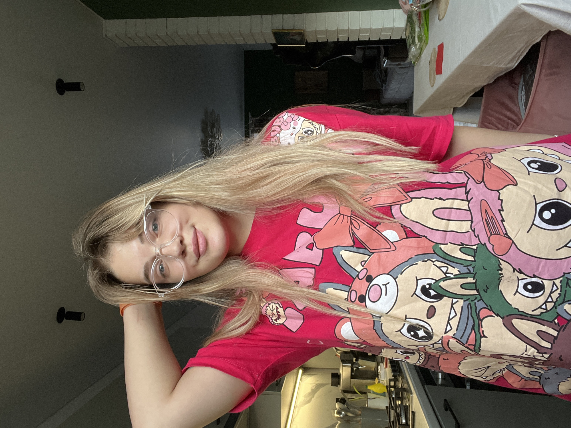 A young woman with long, light blonde hair and clear-framed glasses is featured in an indoor setting, likely a home in Большая Шильна, Russia. She is wearing a vibrant pink t-shirt adorned with numerous cartoon characters, which appear to be stylized animals such as deer or rabbits, rendered with large eyes, bows, and various expressions in shades of pink, beige, and green.
The woman is posing for the camera, looking directly at it with a soft, friendly smile. Her left arm is bent, with her hand gently resting near her head.
The background suggests a casual home environment during daytime, indicated by the bright, natural lighting. To her right, a kitchen area is visible with a countertop and what appears to be appliances or shelving. Further into the background, elements of a dining or living space can be seen, including a table covered with a pink cloth. On the left wall, two dark, cylindrical light fixtures are mounted. No legible text is visible in the image.