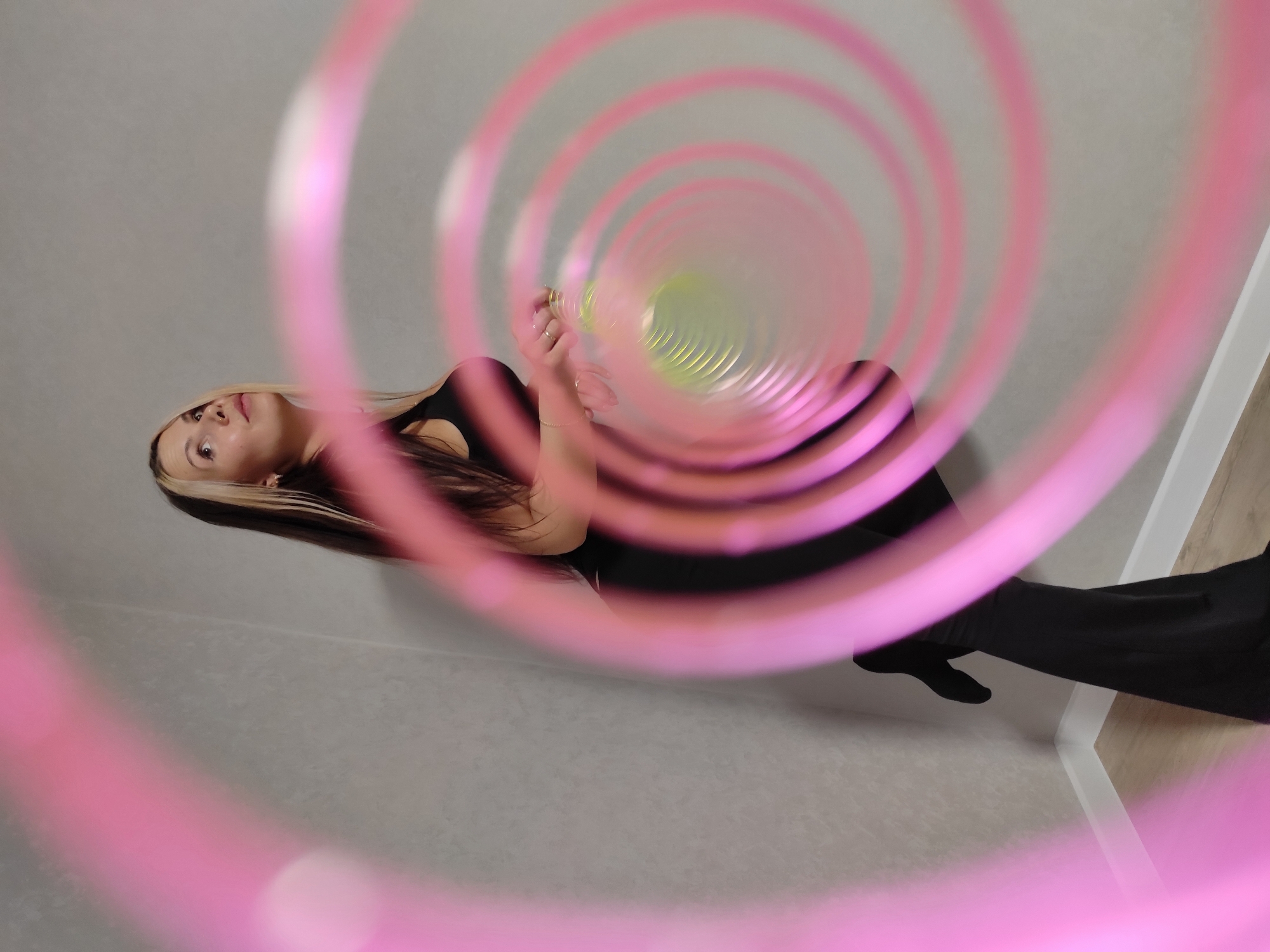 A woman is seen from an unusual, upward perspective, appearing as if she is lying on the floor. She has fair skin, dark hair with blonde streaks, and is wearing a black top and pants. Her arms are raised, and she appears to be interacting with a colorful, spiral-shaped object in front of her. The spiral is predominantly pink and green and occupies the center of the frame, creating a visual tunnel effect. The background consists of a light grey wall and a glimpse of what seems to be a corner with white trim and a light wood-colored floor. The overall lighting is soft and diffuse. There is no explicit indication or detail to definitively identify the location as Chaikovsky, Russia.