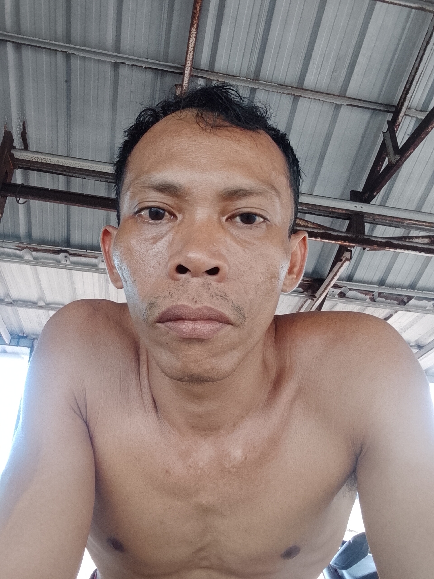 A medium-shot, low-angle photograph depicts a bare-chested man with a weathered complexion. He has short, dark hair and a thin mustache. The man's gaze is directed towards the camera. The background consists of a corrugated metal roof supported by a rusty metal frame. The location is Bukit Ketok, Indonesia.