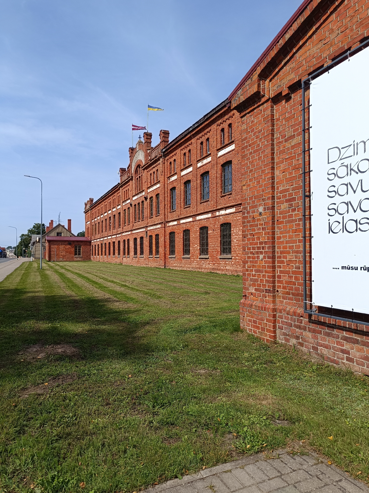 The image shows a red brick building in Aizpute, Latvia. The building is long and has many windows. There are two flags flying from the roof of the building. One is the flag of Latvia and the other is the flag of Ukraine. A large white banner with black text is attached to the side of the building. The building is surrounded by green grass and a sidewalk. A street is visible in the background. The sky is blue and there are some clouds.