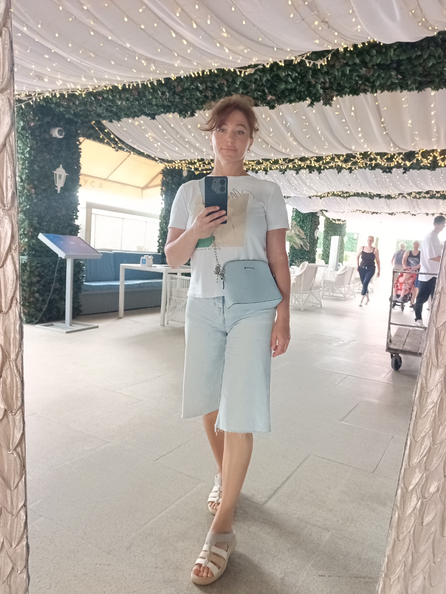 The image shows a woman taking a mirror selfie in what appears to be an outdoor cafe or covered walkway in Sochi, Russia. The woman is wearing a white t-shirt, light blue culottes, and white sandals. She has a light blue crossbody bag. The background shows a reflective surface, likely a mirror, with a decorative frame on the left side. Beyond the mirror, the environment includes lush green wall coverings, draped white fabric with fairy lights, and tables and chairs typical of an outdoor dining area. In the reflection, other people can be seen, including a woman pushing a stroller and another person walking. The flooring is tiled. The overall impression is a bright, possibly evening, setting in a public outdoor space.