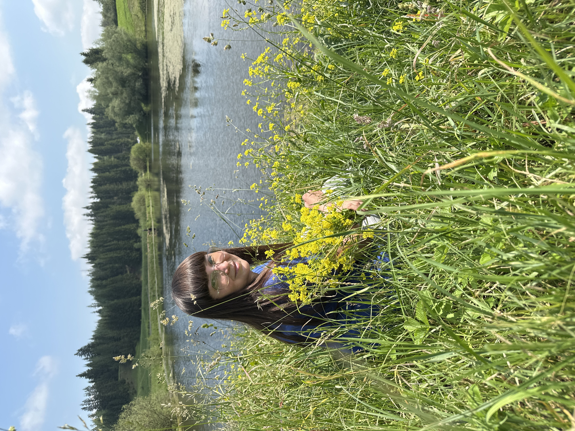 A young female with a fringe and glasses is sitting in tall green grass beside a lake on a sunny day. She is wearing a blue top and holding a bunch of yellow flowers. The lake is surrounded by trees and a green hillside. The sky is blue with a few white clouds.