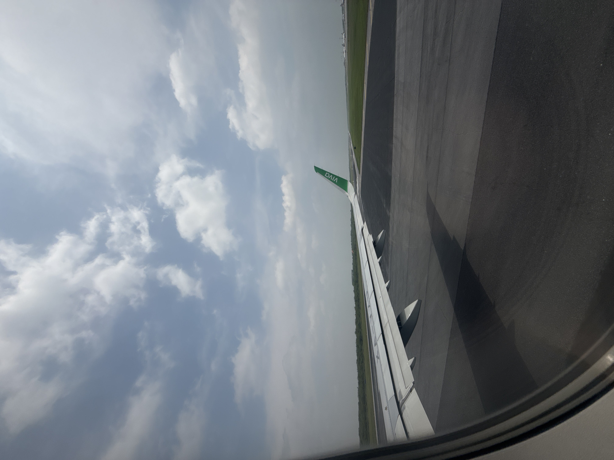 This image captures a view from an airplane passenger window, oriented vertically, likely taken during ground operations at an airport. The aircraft's white wing, featuring a distinct green wingtip with the text "VIVA" in white capital letters, extends diagonally across the middle of the frame. Portions of the airplane's engine nacelles are visible beneath the wing.
To the left of the wing, the sky is partly cloudy, displaying patches of light blue interspersed with white cumulus clouds. The natural lighting suggests it is daytime, possibly late morning or early afternoon, with bright but not harsh illumination.
Below and to the right of the wing, a dark asphalt runway is visible, showing some lighter markings. This paved surface is bordered by a vibrant green grassy area, suggesting the aircraft is either taxiing, awaiting takeoff, or has just landed. The curved frame of the airplane window is partially visible in the bottom right corner of the image.
There are no people visible in the scene. Given the location context of Apodaca, Mexico, the airport is likely Monterrey International Airport (MMTY), which serves as a hub for Viva Aerobus. The image conveys a sense of travel and the commencement or conclusion of a flight.