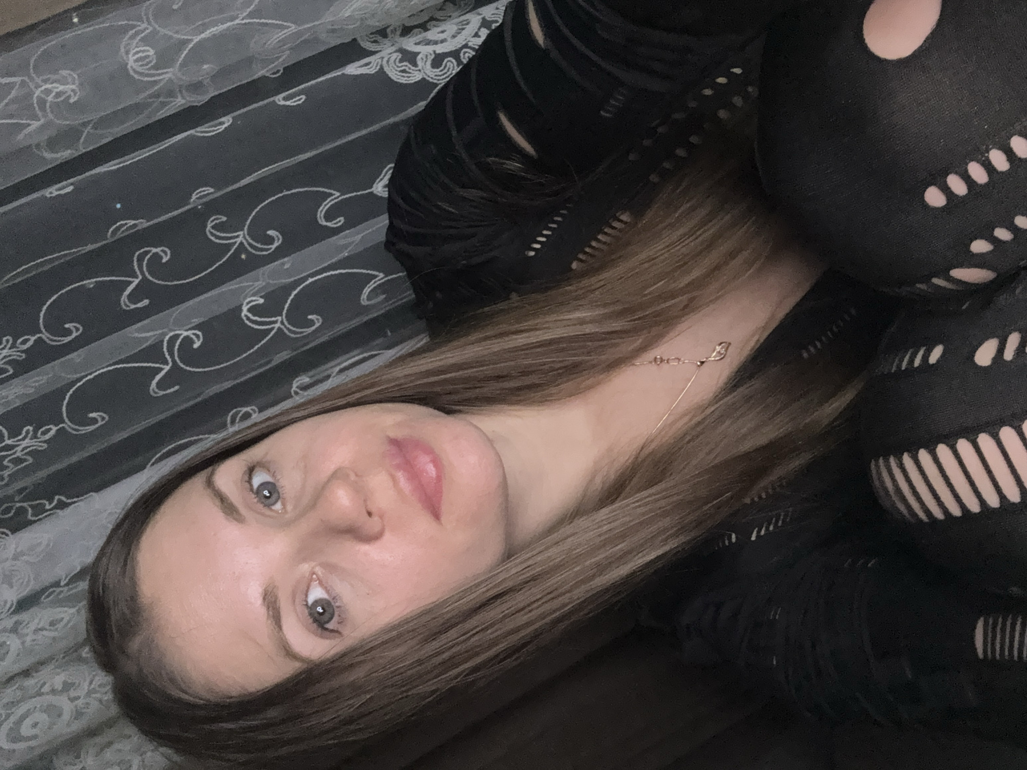A woman with light brown hair is indoors. She is wearing a black mesh top and patterned leggings. She has gray eyes and pink lipstick. The background includes a sheer white curtain with a floral pattern. It is impossible to determine if the location is Batumi, Georgia, or any city without additional information.