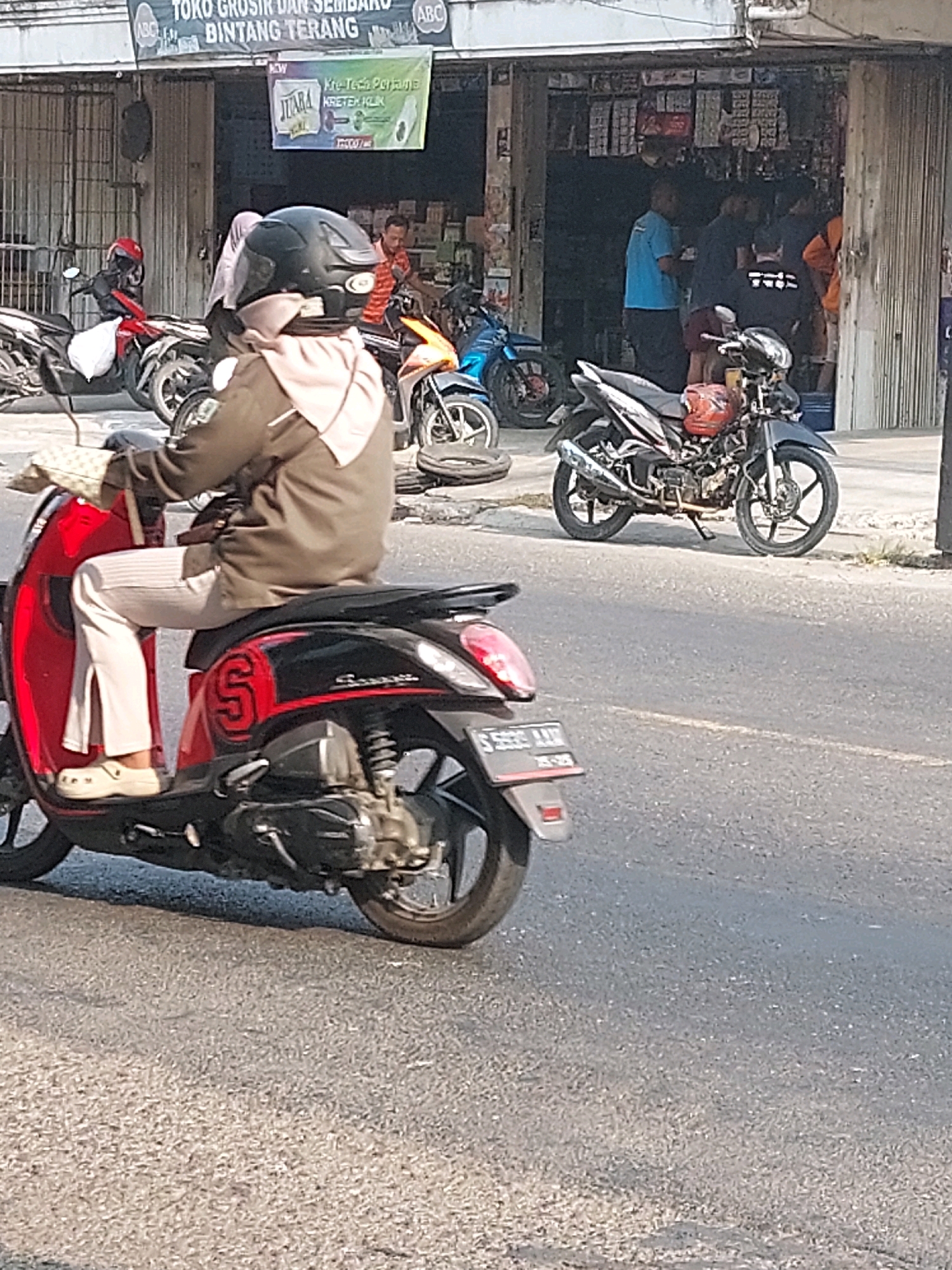 The image depicts a street scene in Sumuragung, Indonesia, on a bright, sunny day. In the foreground, a person wearing a brown jacket, a light-colored hijab, and light-colored pants rides a red and black scooter. The scooter has the letter 'S' prominently displayed on its side, along with the brand name 'Yamaha'. The rider is holding a beige purse. Behind the scooter, a black motorcycle is parked, and further back, a red motorcycle is visible.
The background shows a row of shops with open fronts, displaying various goods. People are seen inside and around the shops. One person in a blue shirt is standing outside one of the shops, while others are partially visible within the storefronts. A banner advertising "JUARA KRETEK KLIK" is hanging on one of the shop fronts. The overall environment suggests a commercial area with moderate pedestrian and vehicle activity. The road surface is asphalt, and there is some grass and vegetation along the edge of the road.