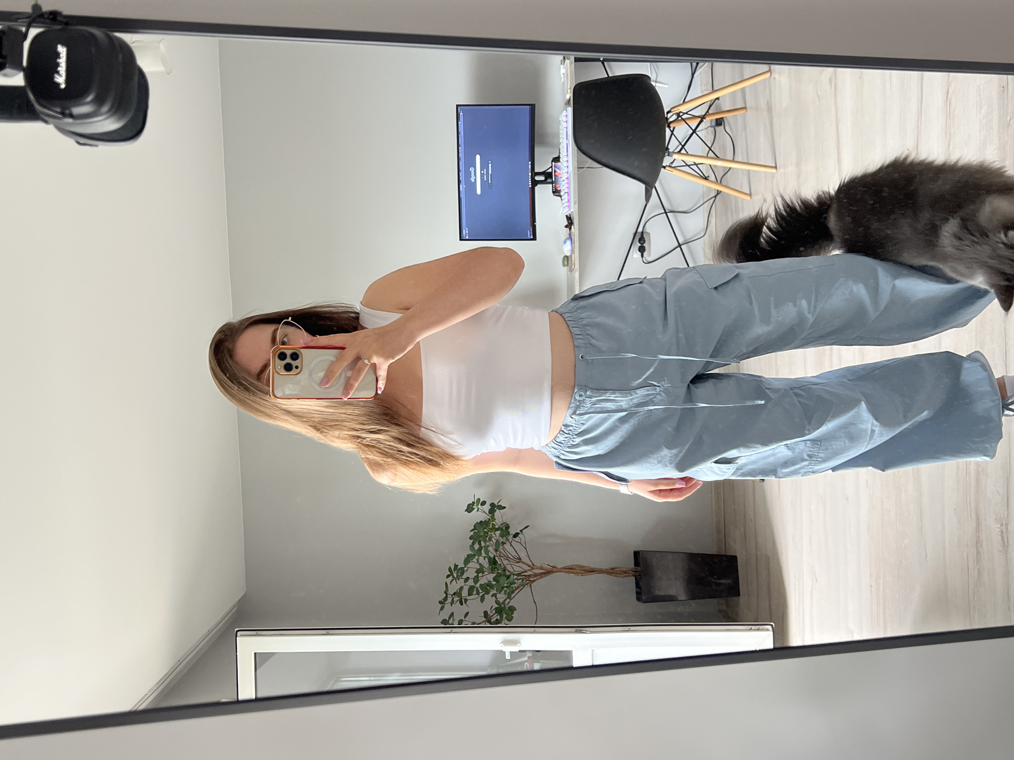 A person is standing in front of a mirror, taking a selfie. They are wearing a white tank top and light blue cargo pants. A cat is visible in the lower right corner of the frame, rubbing against the person's leg. In the background, a computer monitor is on a desk, with a black chair behind it. There is also a plant in a pot to the left of the person. The scene appears to be indoors, possibly in an apartment. The location in Ternopil, Ukraine is not discernible from the image.