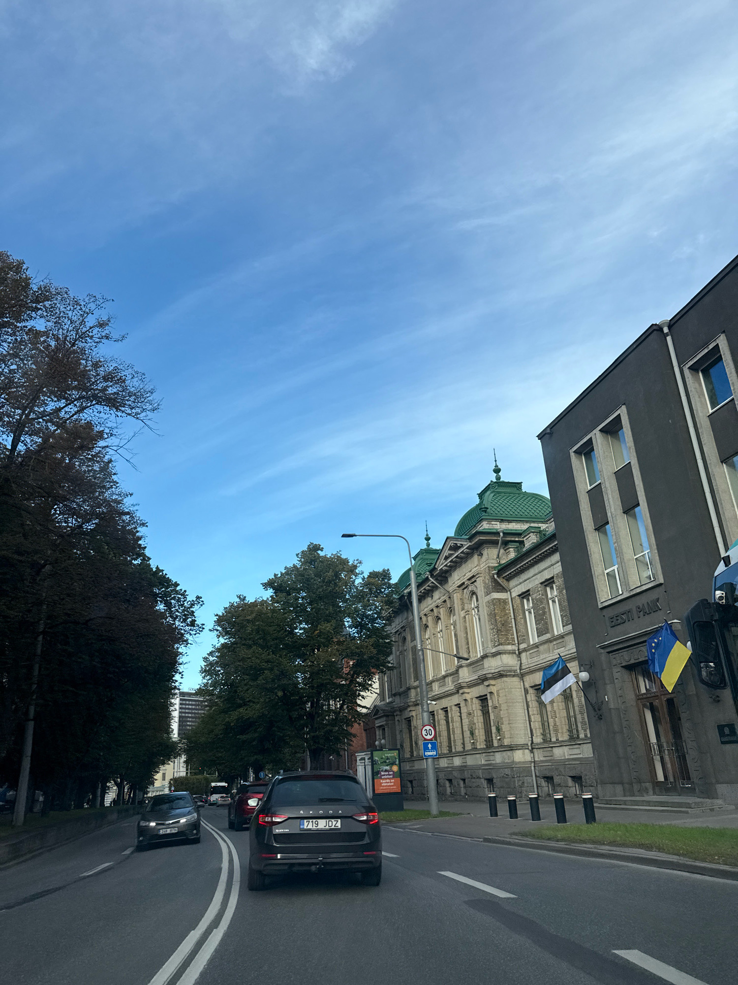 The image shows a view from a vehicle driving on a street in Tallinn, Estonia, during daylight hours under a partially cloudy blue sky. On the right side of the street, there are two prominent buildings. The building in the foreground is a modern, dark-colored structure with rectangular windows, featuring the text "EESTI PANK" above its entrance. An Estonian flag and a Ukrainian flag are displayed on its facade. Behind this building, to the left, is an older, ornate building with a distinctive green dome and arched windows. Trees line the left side of the street, with some showing autumnal colors. Several cars are visible on the road, including a dark-colored Skoda in the foreground. A street lamp and traffic signs are also present. The scene suggests a normal day with vehicular traffic in an urban environment.