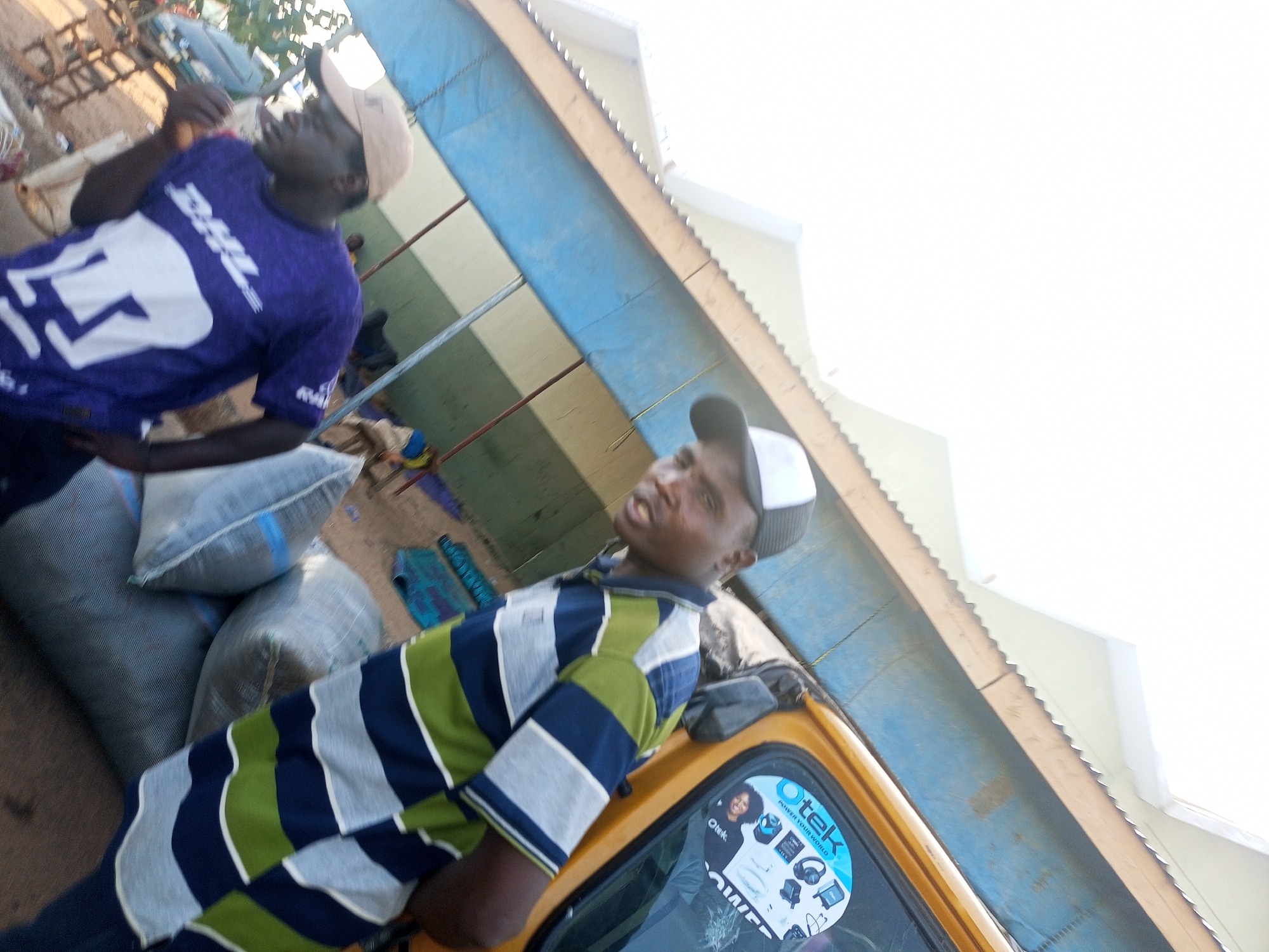 A candid, slightly angled shot depicts two men alongside a yellow vehicle. One man is clad in a striped shirt and a baseball cap, leaning against the vehicle with a sticker on the window featuring the word "Otek". Adjacent to them are large sacks, presumably contents from the vehicle. Behind and to the left, another man is wearing a purple shirt with the letters "DHL" emblazoned on it and is sporting a baseball cap as well. The surrounding area is vaguely discernable and appears to be outdoors. The location is in the city of Unknown, Unknown.