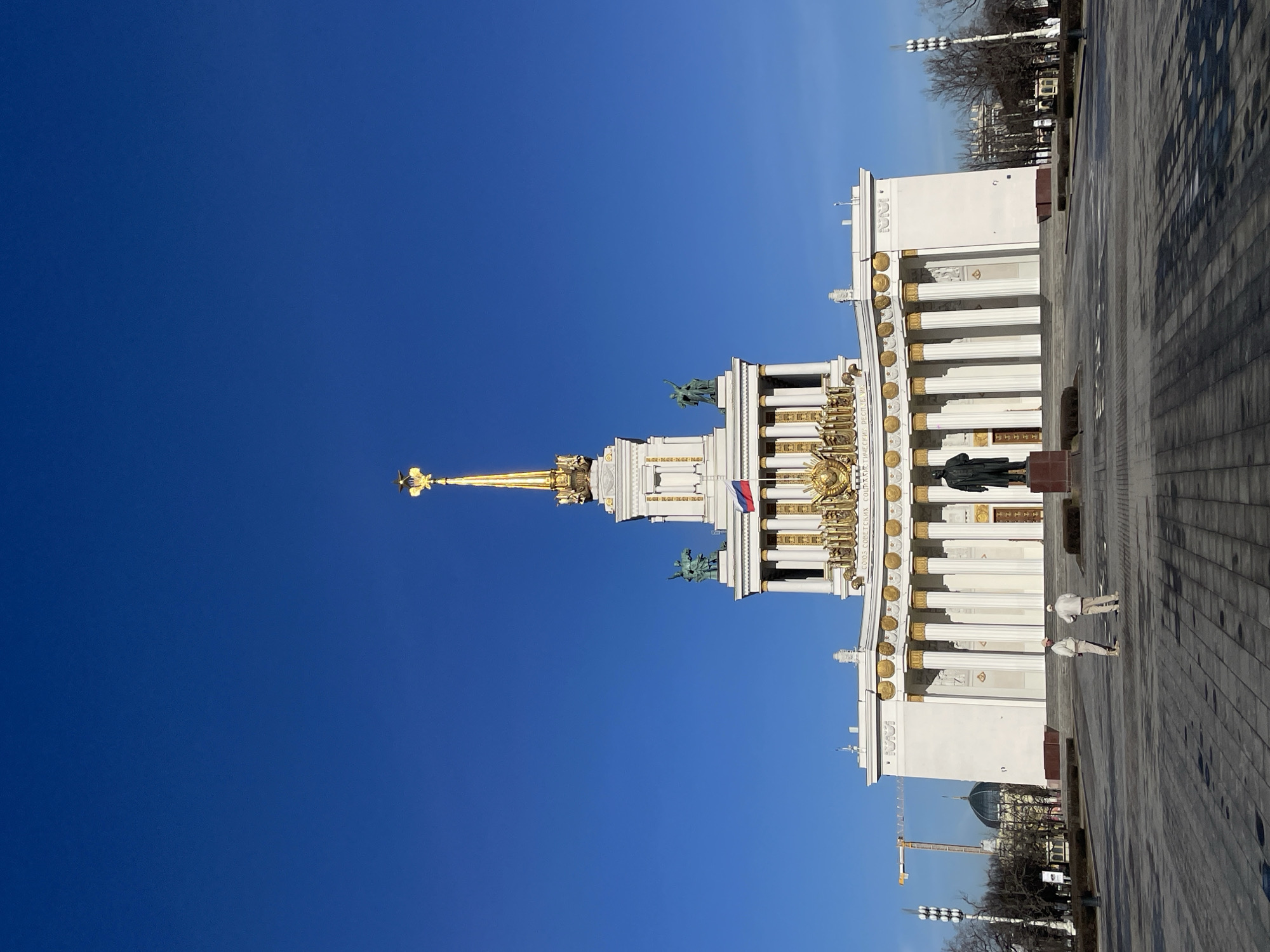 The image shows a view of the Central Pavilion in the VDNKh park in Moscow, Russia. The building features a white facade with golden decorative elements, including columns and statues. A tall golden spire stands prominently on top of the pavilion. A Russian flag is visible. In front of the pavilion, there is a statue and two people walking on a paved area. The background includes a clear blue sky. A construction crane and a dome-shaped building can be seen in the distance. The photograph is taken on a sunny day with clear visibility.