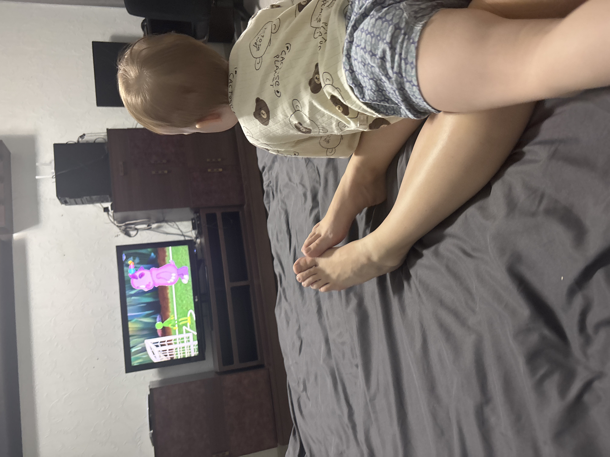 A child sits on a gray bed in a room with a television showing a cartoon. The room contains a dark-colored entertainment center with dark-colored electronics. The child is wearing pajamas with a brown bear pattern and blue shorts. The location of Ферма, Russia is not discernible from the image.