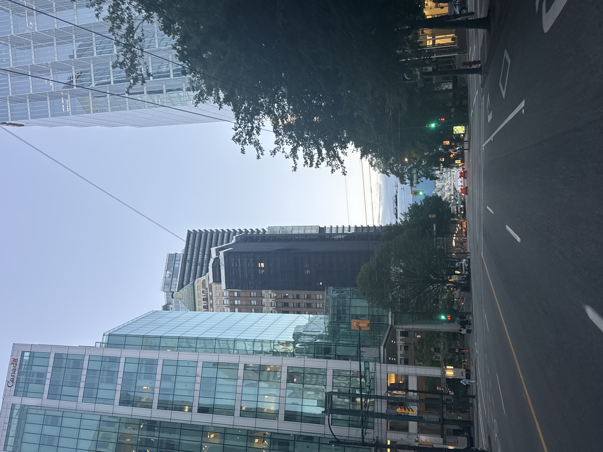 The image shows a street view in downtown Vancouver, Canada. The scene is captured from a vehicle's perspective, looking down a wide city street lined with modern glass-fronted skyscrapers on both sides. One prominent building on the left has a "Canada" logo at the top. Traffic lights are visible, with one showing green. Trees line the sidewalks, and street signs indicate "W Pender St." Pedestrians can be seen on the sidewalks and crossing the street. In the distance, a body of water and mountains are visible, suggesting a waterfront area of the city. The sky is a pale blue, indicating it might be early morning or late afternoon. The overall impression is of a bustling urban environment.