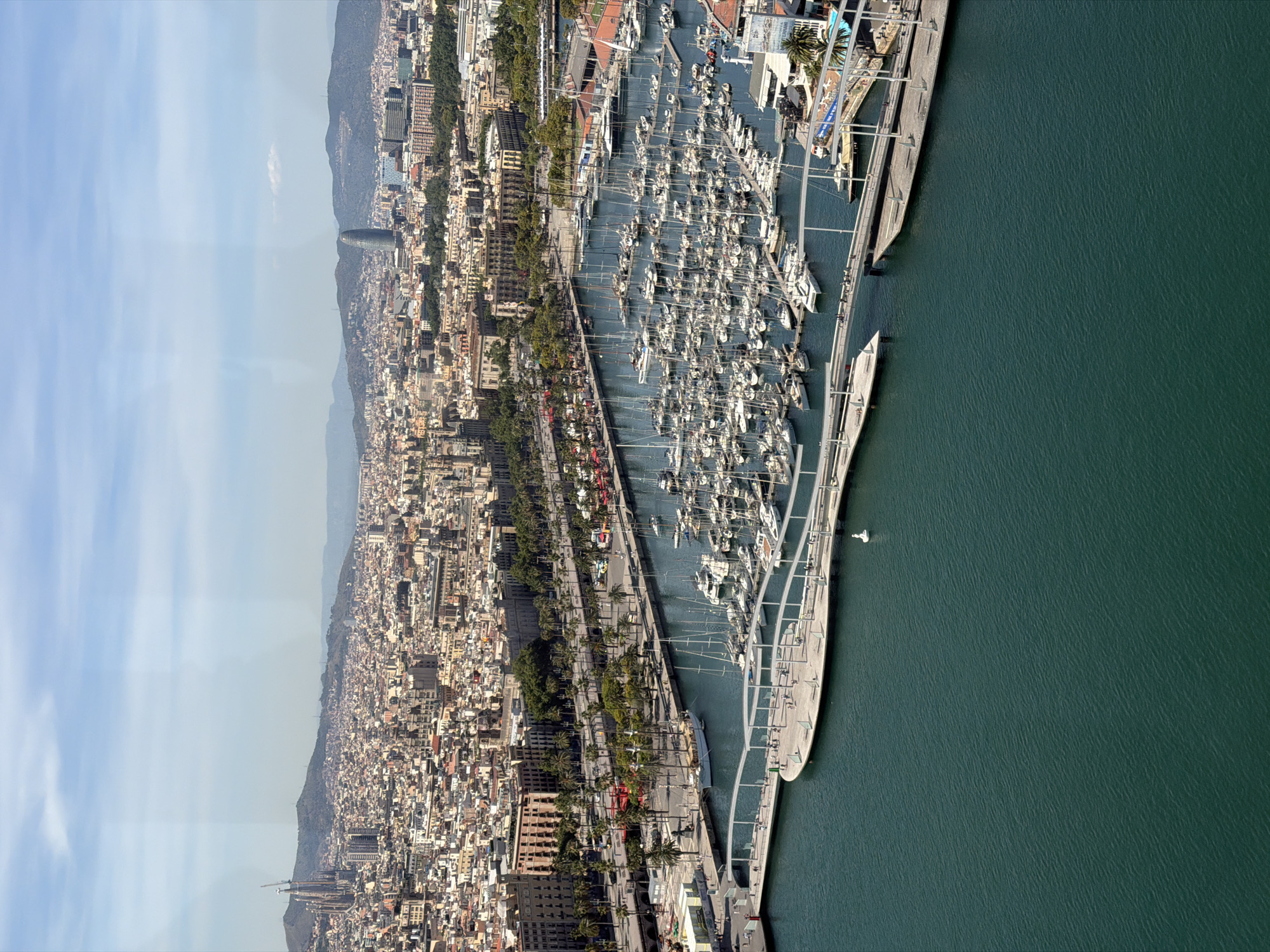 The image is an aerial view of the Port Vell marina in Barcelona, Spain. The marina is filled with numerous sailboats and yachts, docked closely together. A curved, elevated walkway extends over the water, with people visible walking on it. Behind the marina, a bustling cityscape stretches out, characterized by dense urban development with numerous buildings of varying heights and architectural styles. In the distance, a range of hills forms a natural backdrop to the city. One prominent skyscraper, the Agbar Tower, is visible among the buildings, and further in the background, the spires of the Sagrada Familia church can be seen on a distant hill. The sky is mostly clear with scattered clouds, and the water in the marina is a deep teal color. The scene captures a vibrant mix of maritime activity and urban life.