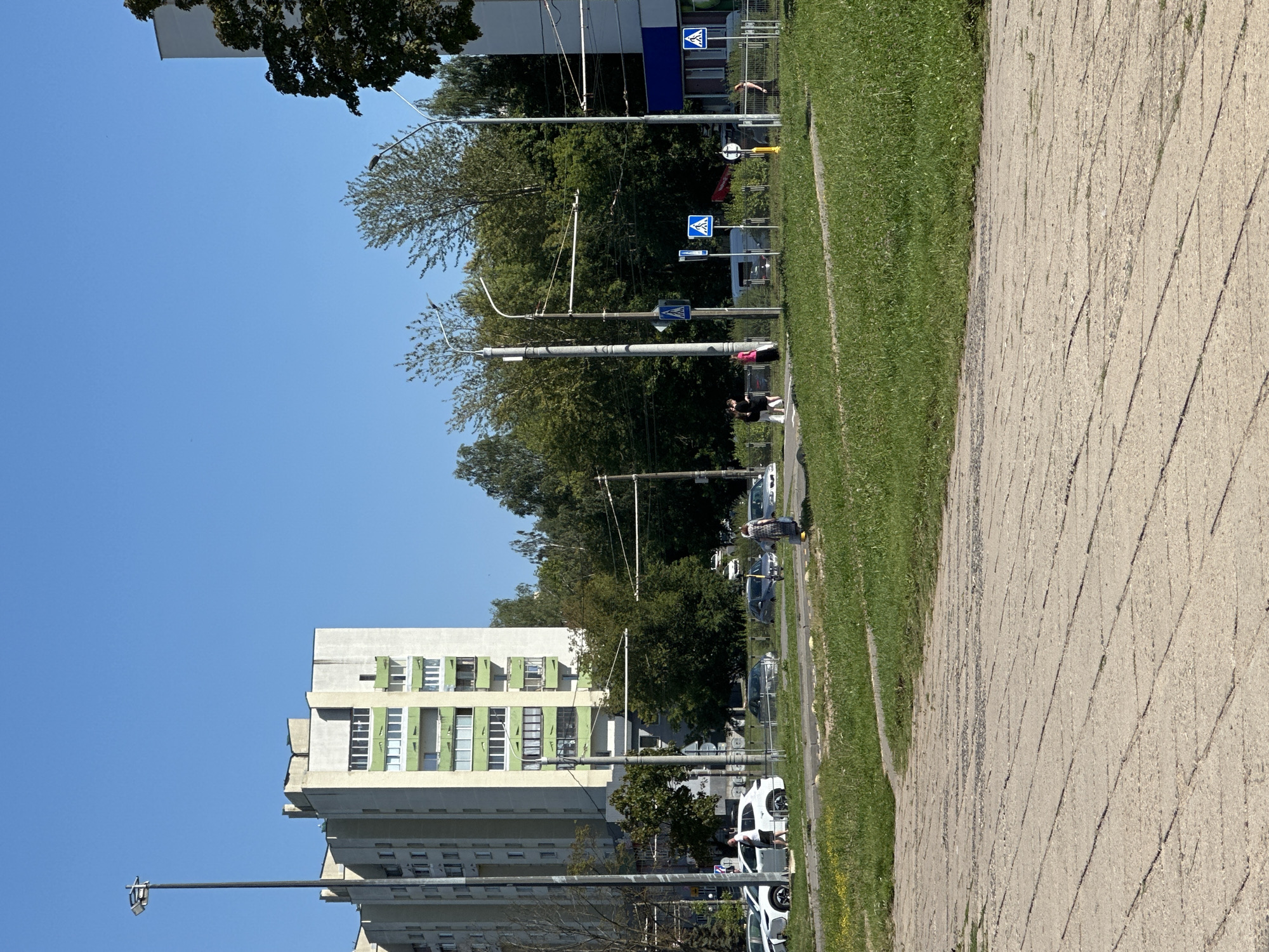 This is an outdoor, eye-level, medium shot captured on a sunny day, showcasing an urban scene in Minsk, Belarus. The foreground features a cracked concrete pathway transitioning into a manicured green lawn. Behind the lawn, a street is visible with several parked cars and pedestrians. A tall, light-colored apartment building with green accents on its balconies dominates the left side of the frame. Trees line the background, interspersed with streetlights and traffic signals. A white car is parked on the left, and two individuals are standing next to it. Further to the right, a woman is walking with a yellow shopping bag, and other pedestrians are visible near crosswalks. Power lines and overhead tram wires are also present. The sky is clear and blue.