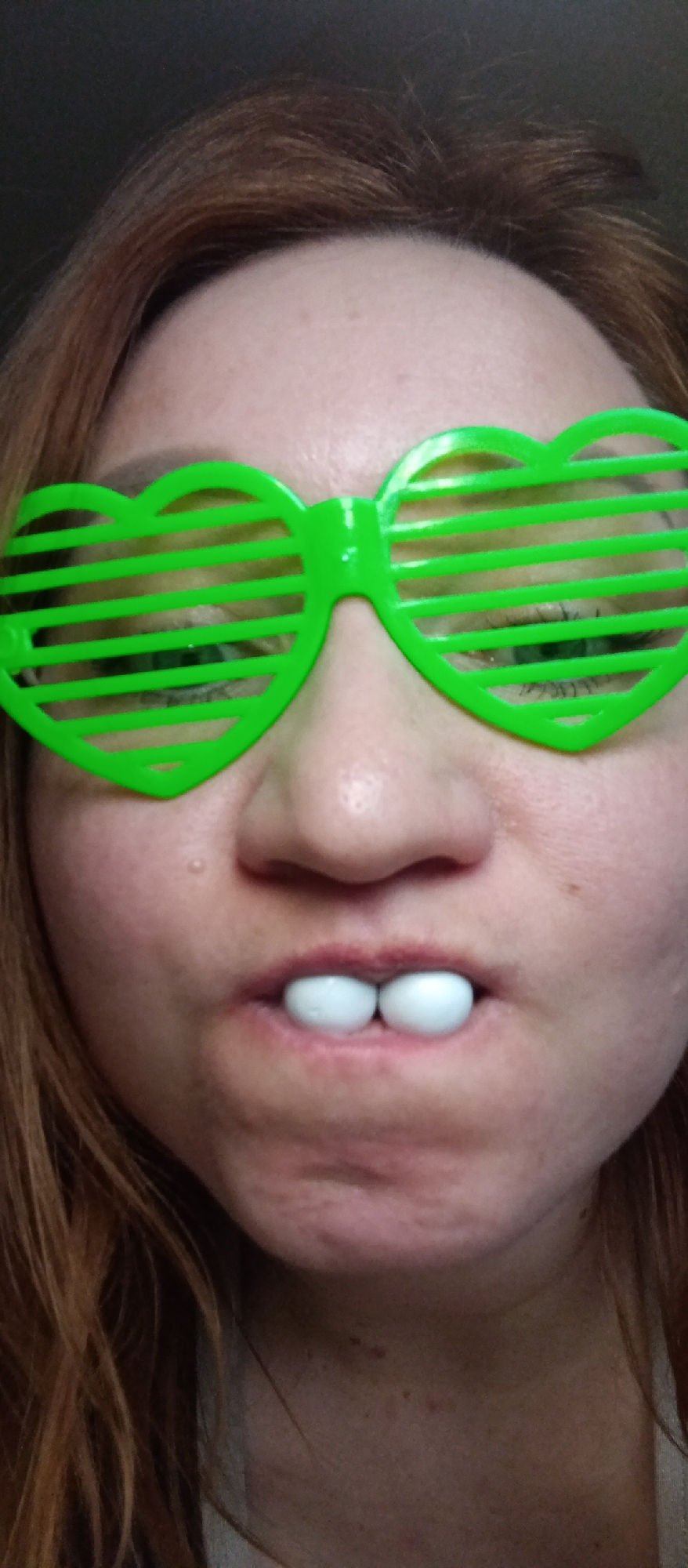 A close-up, headshot-style portrait captures a person, likely female, from their forehead to their upper chest/neck area. The subject has reddish-brown hair, visible around the sides and top of their head, framing a fair-skinned face.
The most prominent features are a pair of bright green, heart-shaped novelty "shutter" glasses worn over the eyes. These glasses feature horizontal slats, through which parts of the subject's eyes are visible. Just below the nose, the subject is holding two small, white, spherical or oval objects (resembling candies or gumballs) between their upper and lower lips, which are slightly parted. The lips have a natural, somewhat reddish hue.
The subject's expression is difficult to fully gauge due to the glasses and items in the mouth, but the overall presentation suggests a playful or humorous intent. Minor skin texture and a few small blemishes are visible on the face, particularly around the cheeks, chin, and upper neck.
The background is dark and out of focus, indicating an indoor setting with minimal distractions. There are no visible location cues or text within the image. The lighting appears even, suggesting an indoor light source.
Location context: Unknown, Unknown