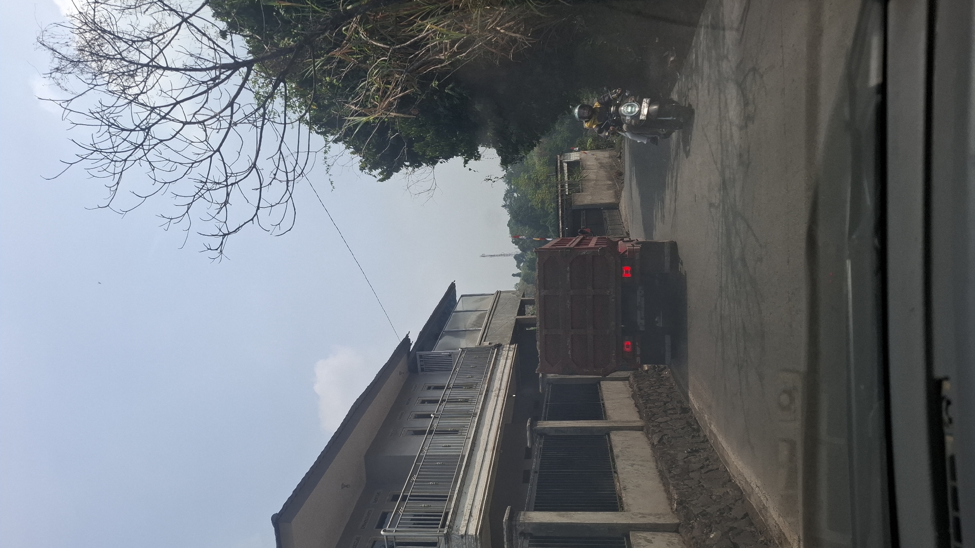 The image depicts a street scene in Sirnagalih, Indonesia, on a clear, bright day. A red flatbed truck is driving down the paved road, with its rear end prominently featured. To the right of the truck, a person on a scooter wearing a yellow jacket and helmet is also traveling in the same direction. On the left side of the road, a multi-story building with a balcony and a fence lines the street. Lush green trees and vegetation are visible on the right side and in the background, with a distant tower also visible. The perspective suggests the image was taken from inside a vehicle.