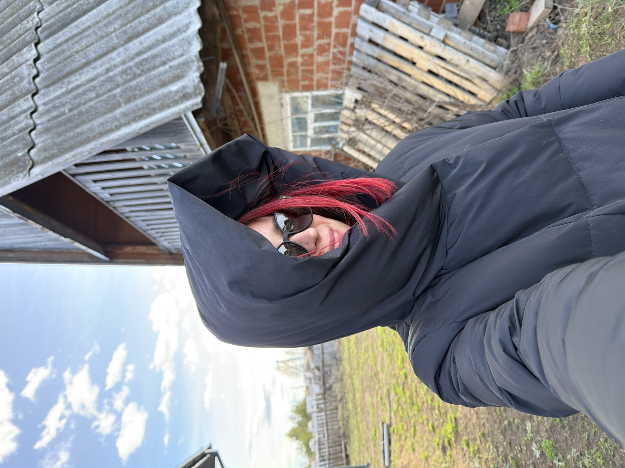 A selfie shows a woman with red hair wearing a black hooded puffer jacket and sunglasses. The background is slightly blurry and includes a dilapidated brick building, a wooden pallet leaning against it, and a shed with a corrugated metal roof. There's also a field with a wooden fence visible in the distance. Based on image metadata, the location is Chaikovsky, Perm Krai, Russia.