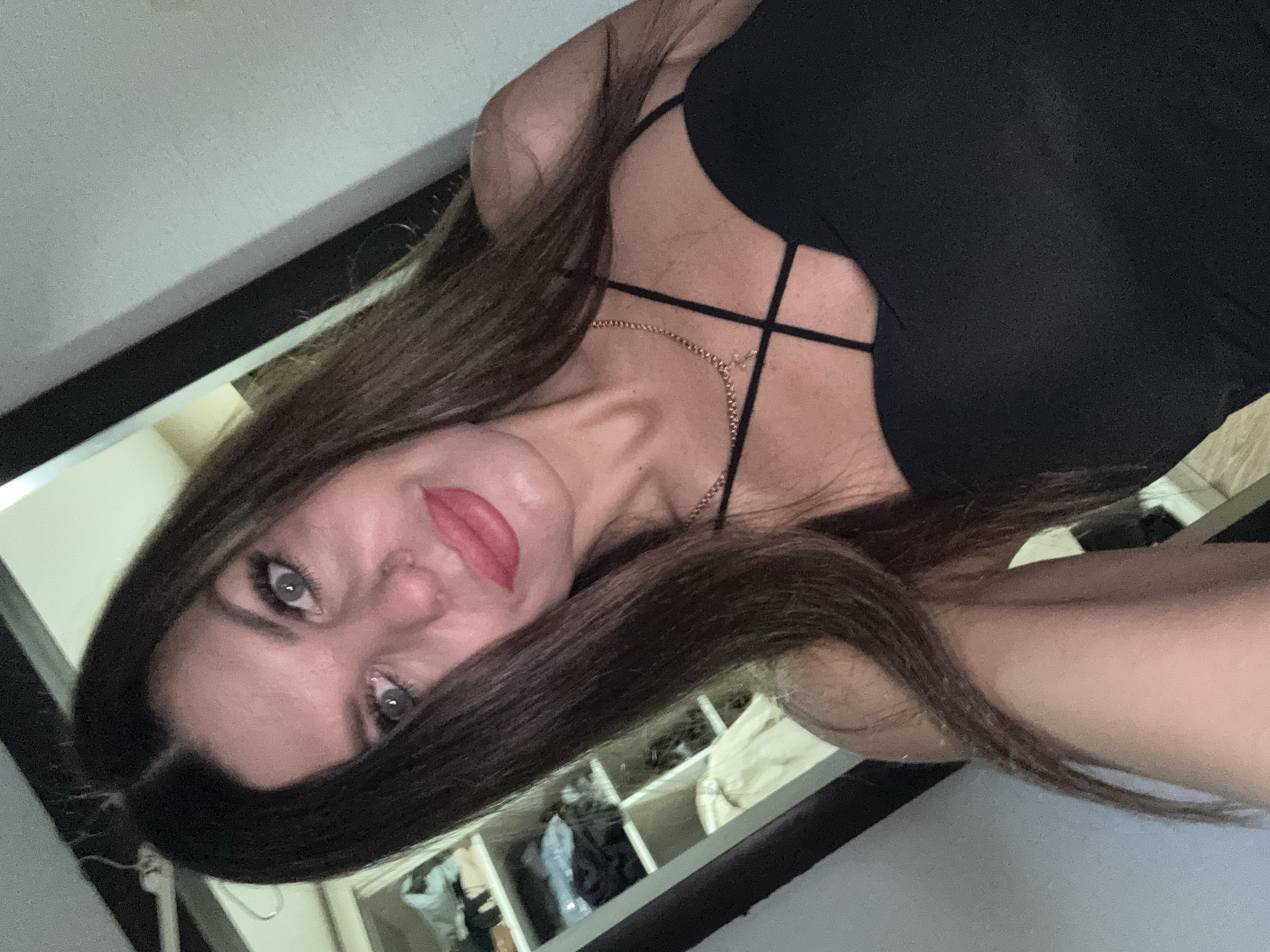 A woman with long, dark brown hair and blue eyes is taking a selfie in what appears to be a bedroom or dressing area. She is wearing a black, strappy top and a delicate gold necklace with a cross pendant. Behind her, a mirror reflects a closet filled with clothes on shelves. The lighting is dim, creating a soft glow. The woman has makeup on, with red lipstick and false eyelashes. The setting is indoors, and the overall impression is one of personal preparation or a casual moment captured. There is no indication of the city of Chaikovsky, Russia, in the image itself.